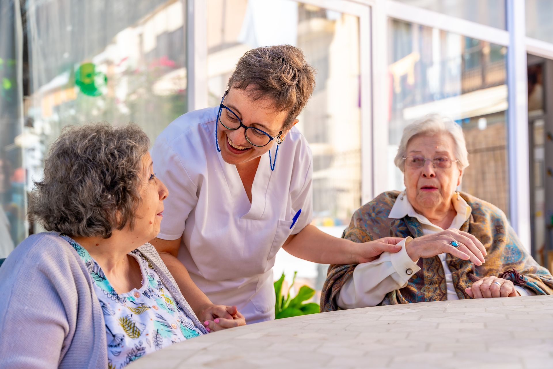 Caregiver in white uniform smiles at two elderly women seated outdoors.