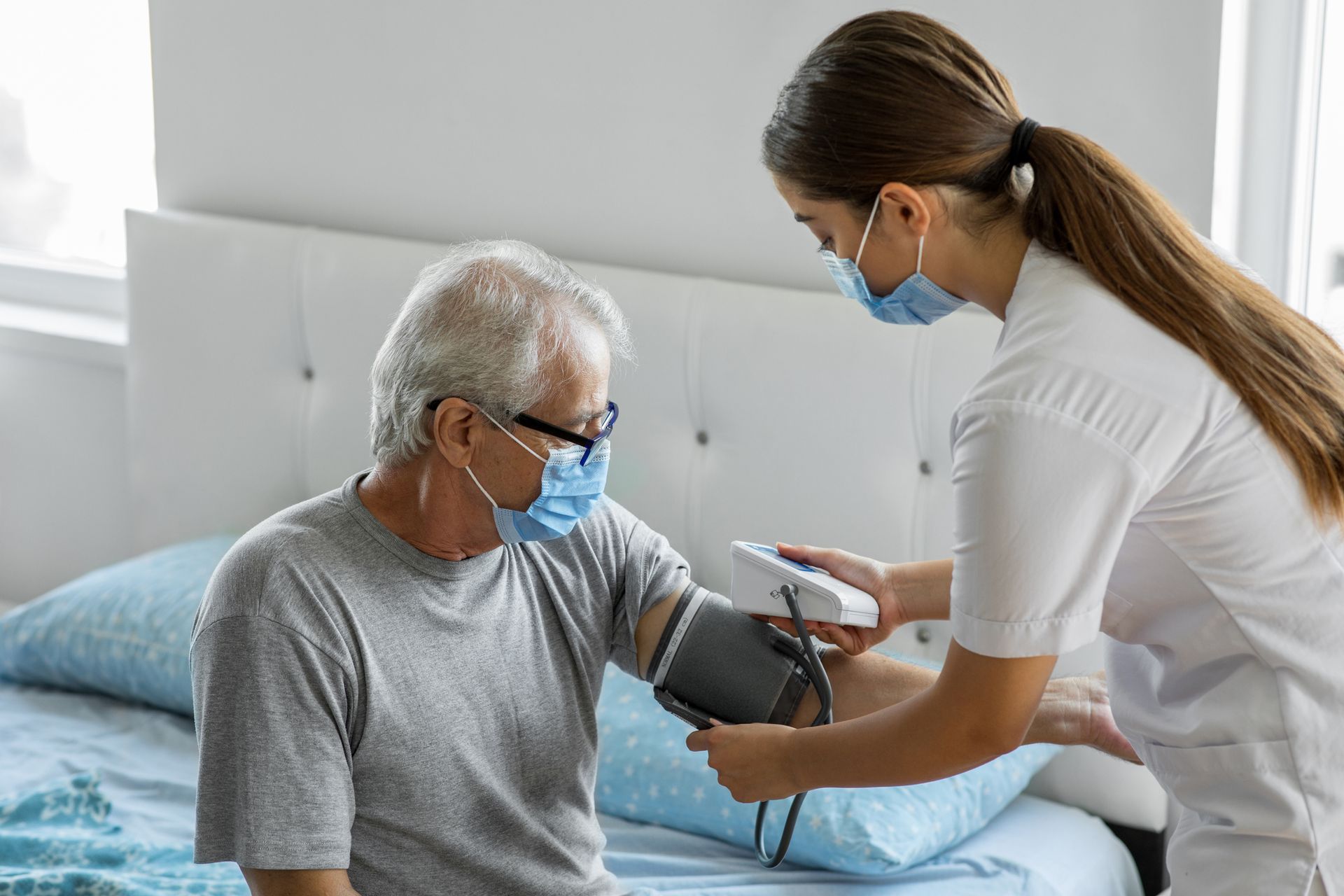 Nurse taking an elderly man's blood pressure in a bedroom, both wearing masks.