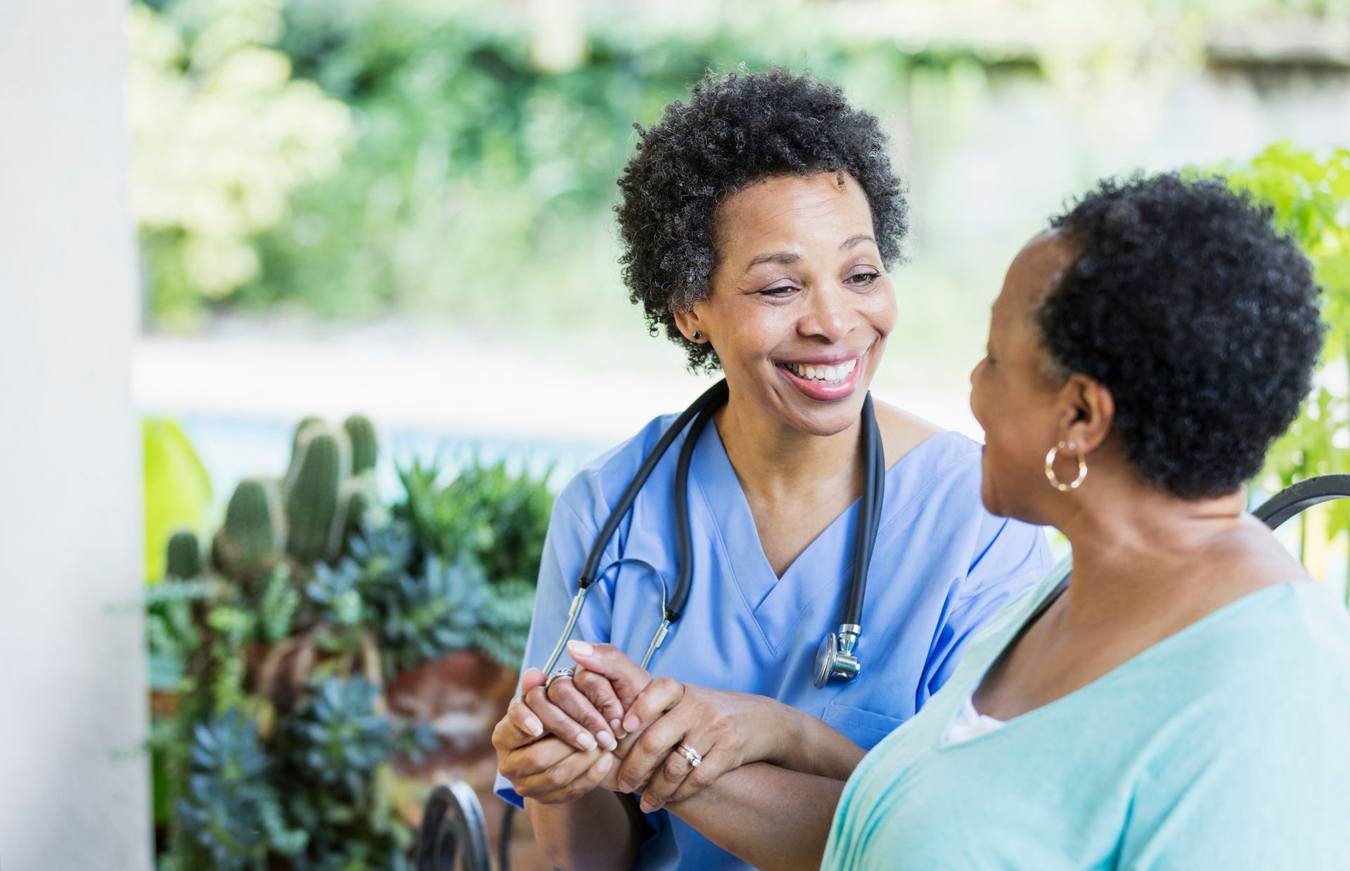 A smiling healthcare worker in blue scrubs holds the hand of an older Black woman, both outdoors.