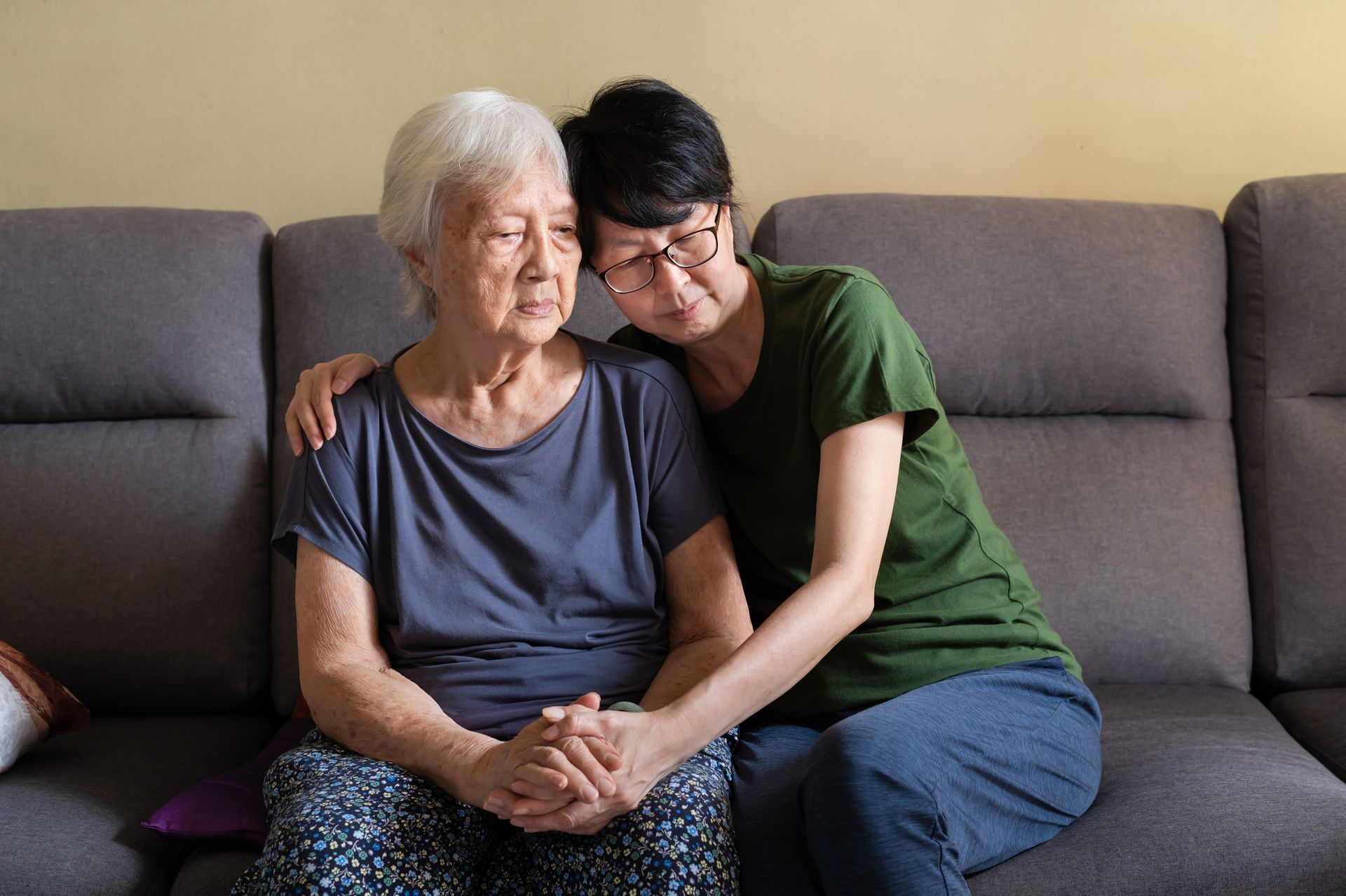 Woman comforts elderly woman on a sofa. Both are Asian; one is hugging and holding the other's hand. Sad expressions.