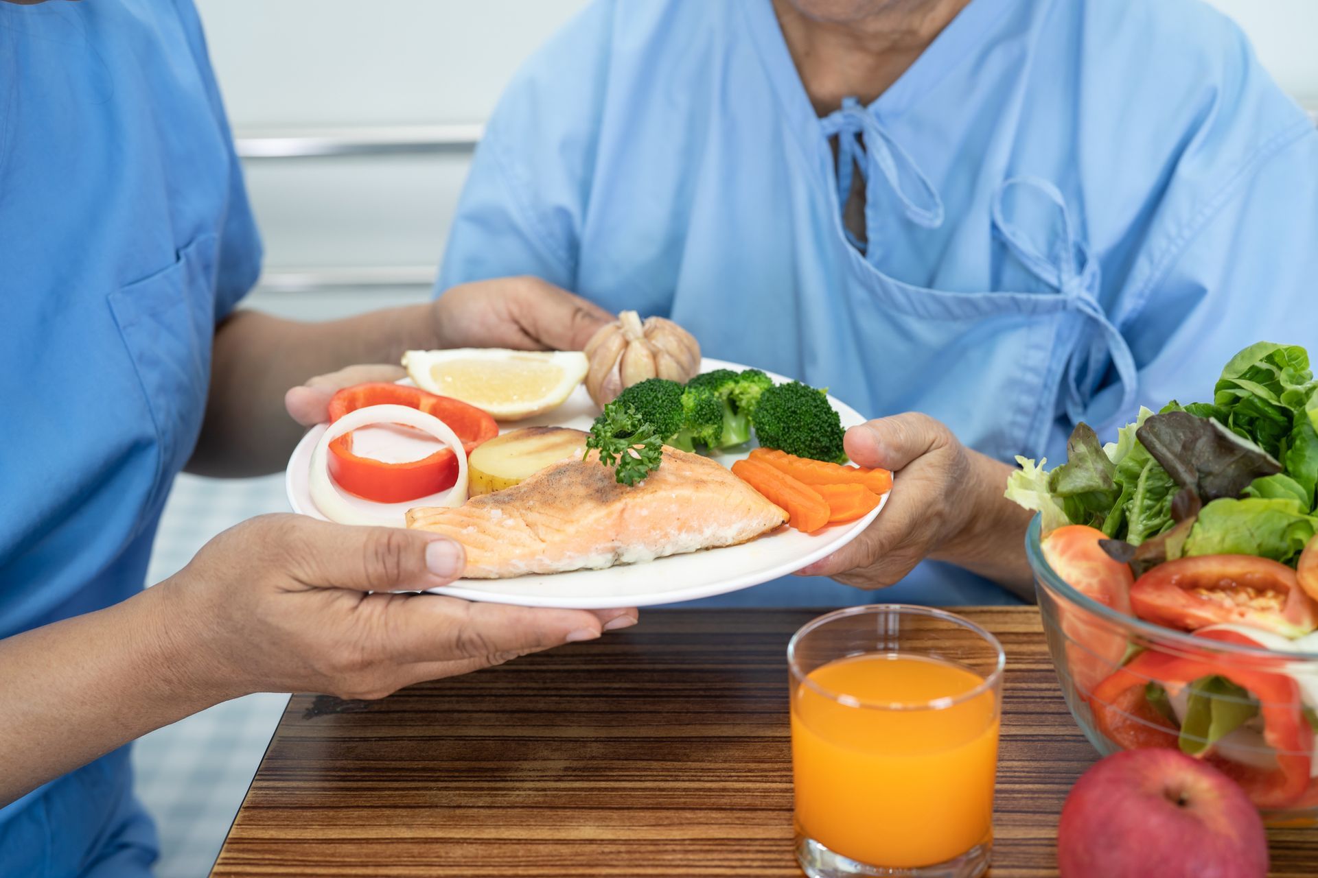 Caregiver serving a plate of food (salmon, vegetables) to a patient in a hospital.