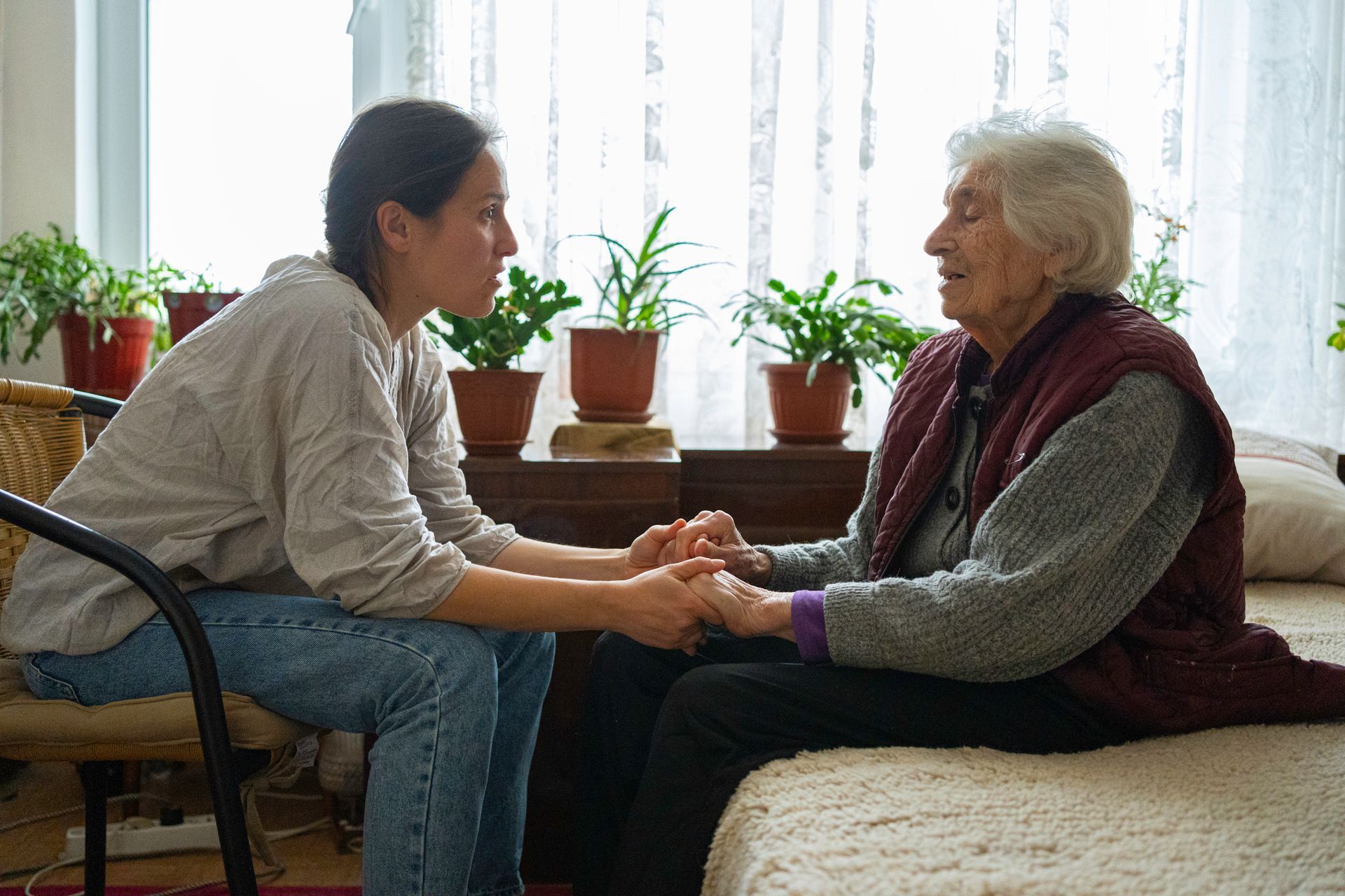 Young woman holds hands with elderly woman indoors. Both are looking at each other, engaged in conversation.