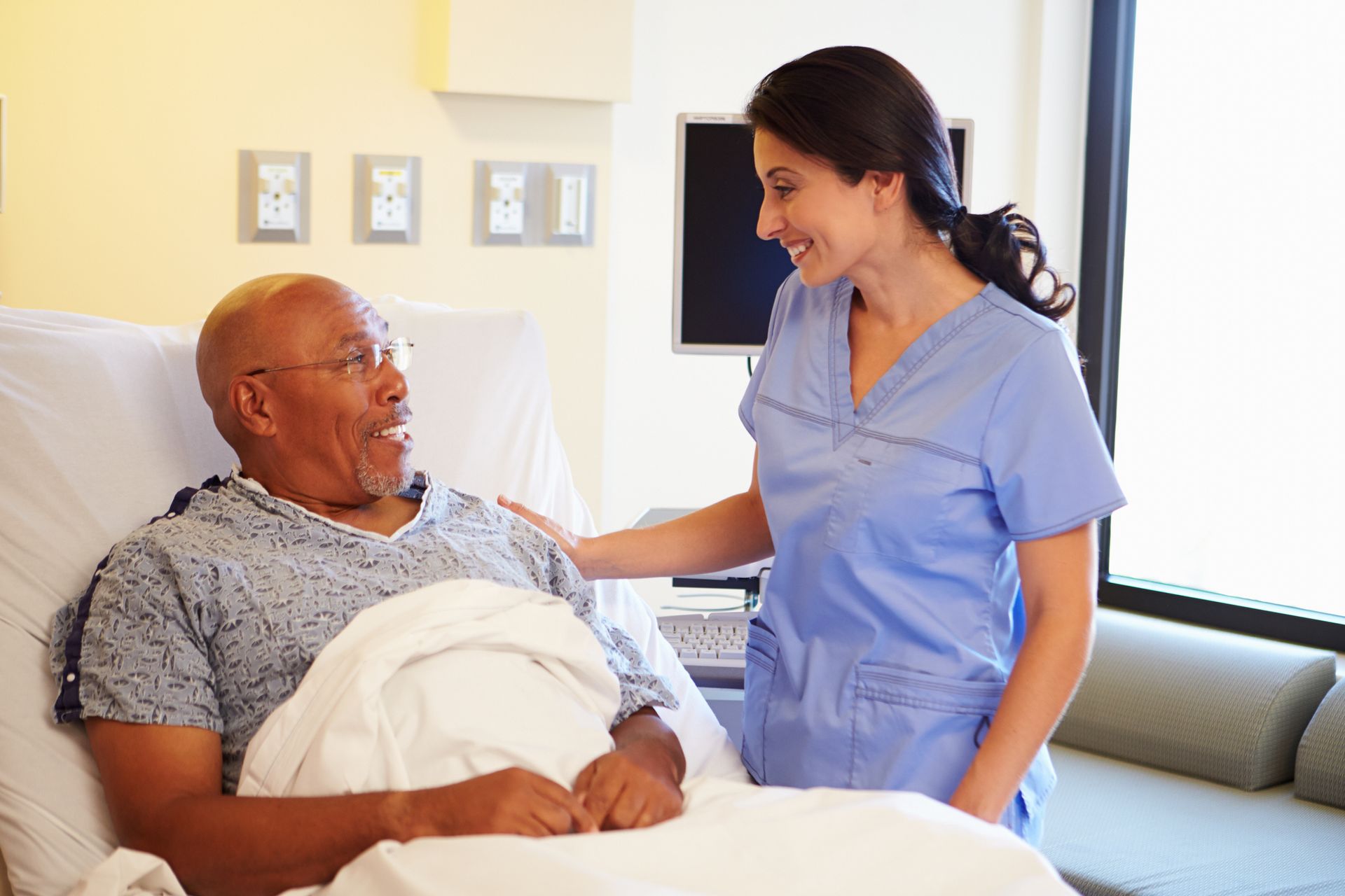 Nurse in blue scrubs smiles, placing a hand on shoulder of elderly Black man in hospital bed.
