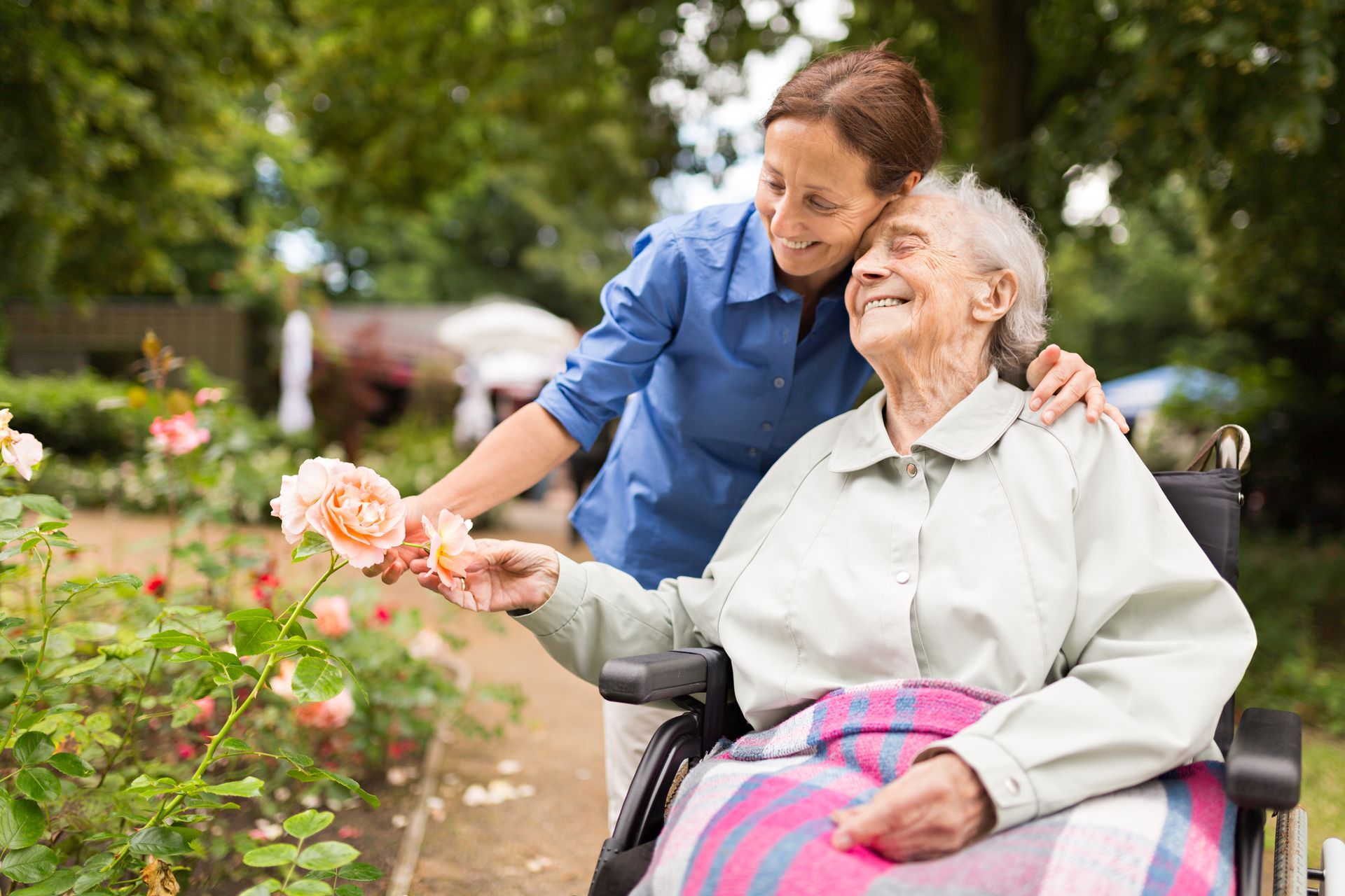 Woman in wheelchair smiles at flowers with the assistance of a caregiver in a garden.