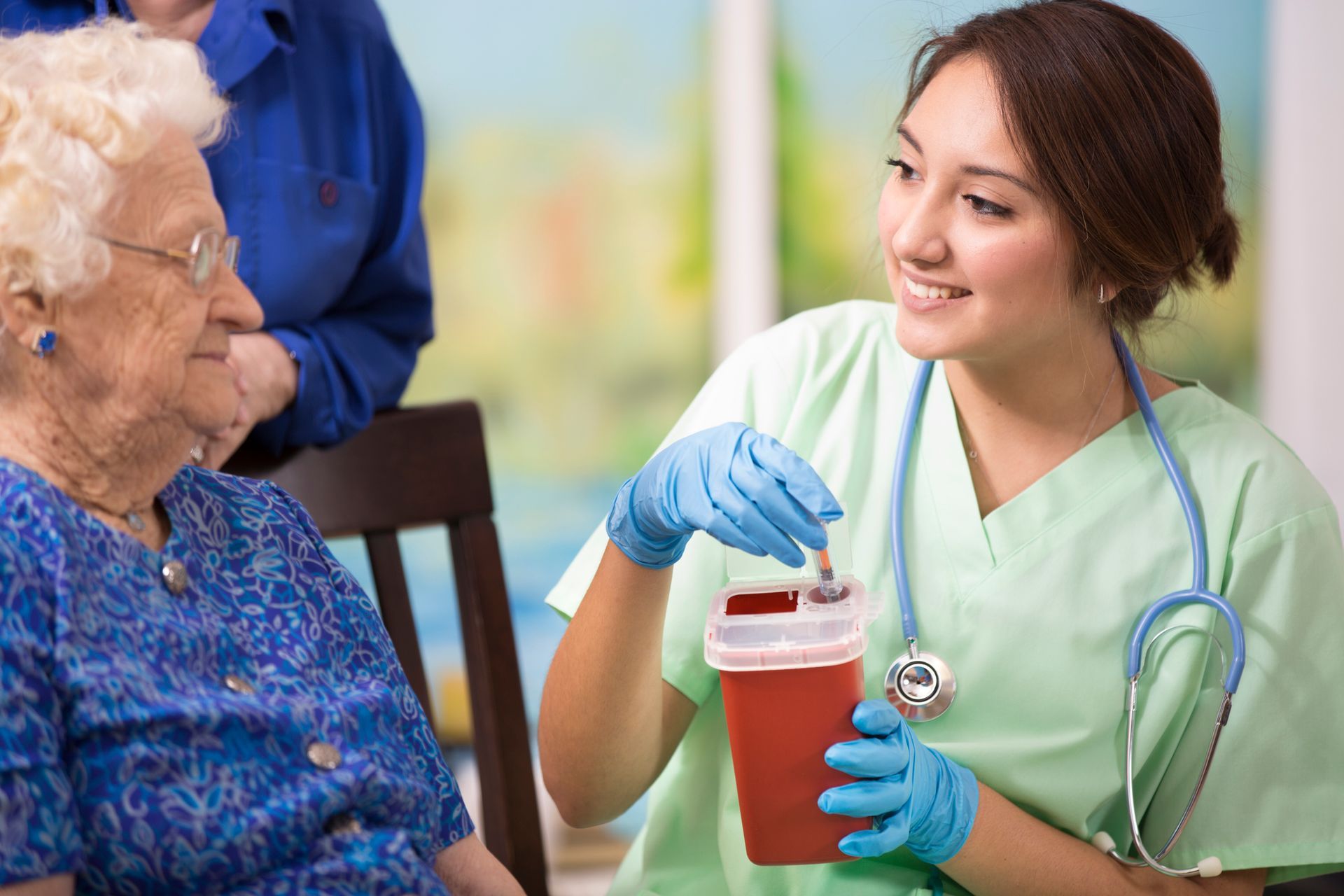 Nurse preparing a syringe, talking to an elderly woman; a second woman looks on. Indoor setting.