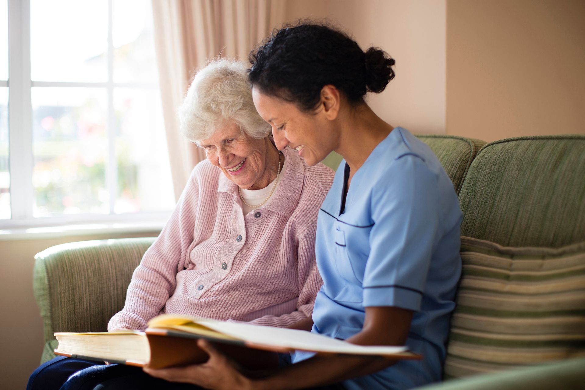 Woman caregiver and senior woman on sofa, looking at a book. Both smile.