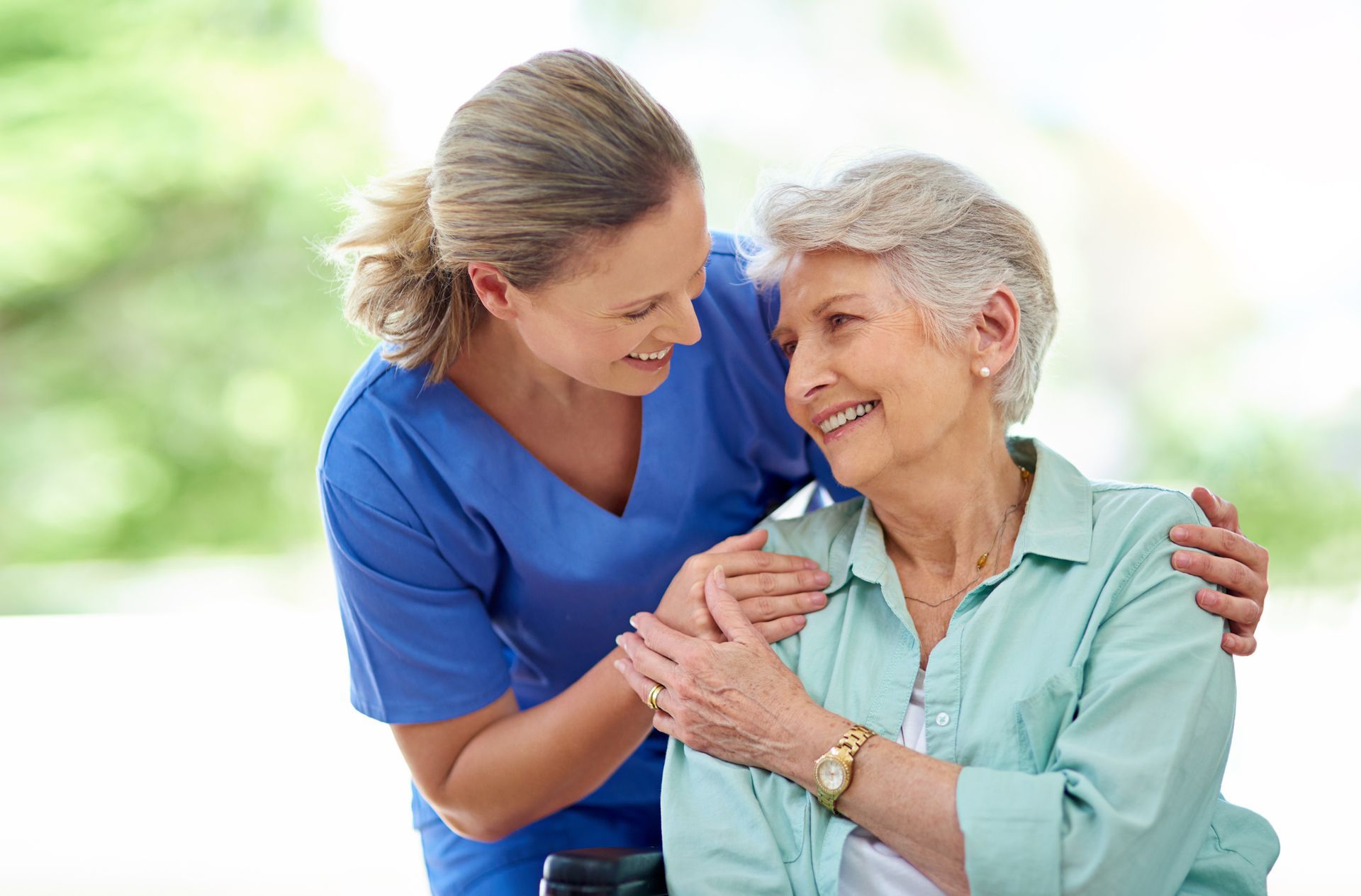 Caregiver in blue scrubs embraces elderly woman with gray hair, smiling. Outdoors, bright.
