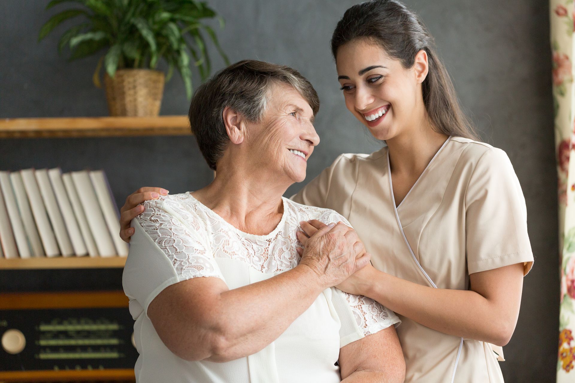 Woman caregiver with arm around elderly woman, both smiling. Interior setting, bookshelves.