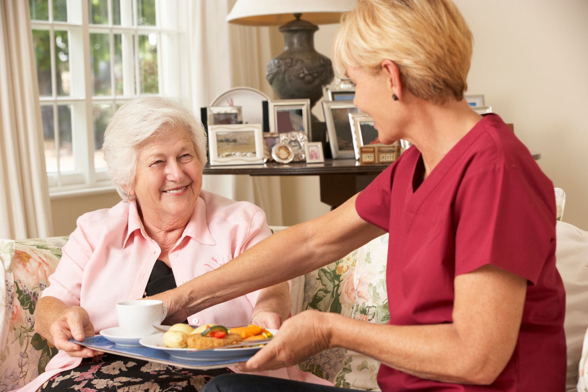Caregiver serving food to an elderly woman at home; both smiling.