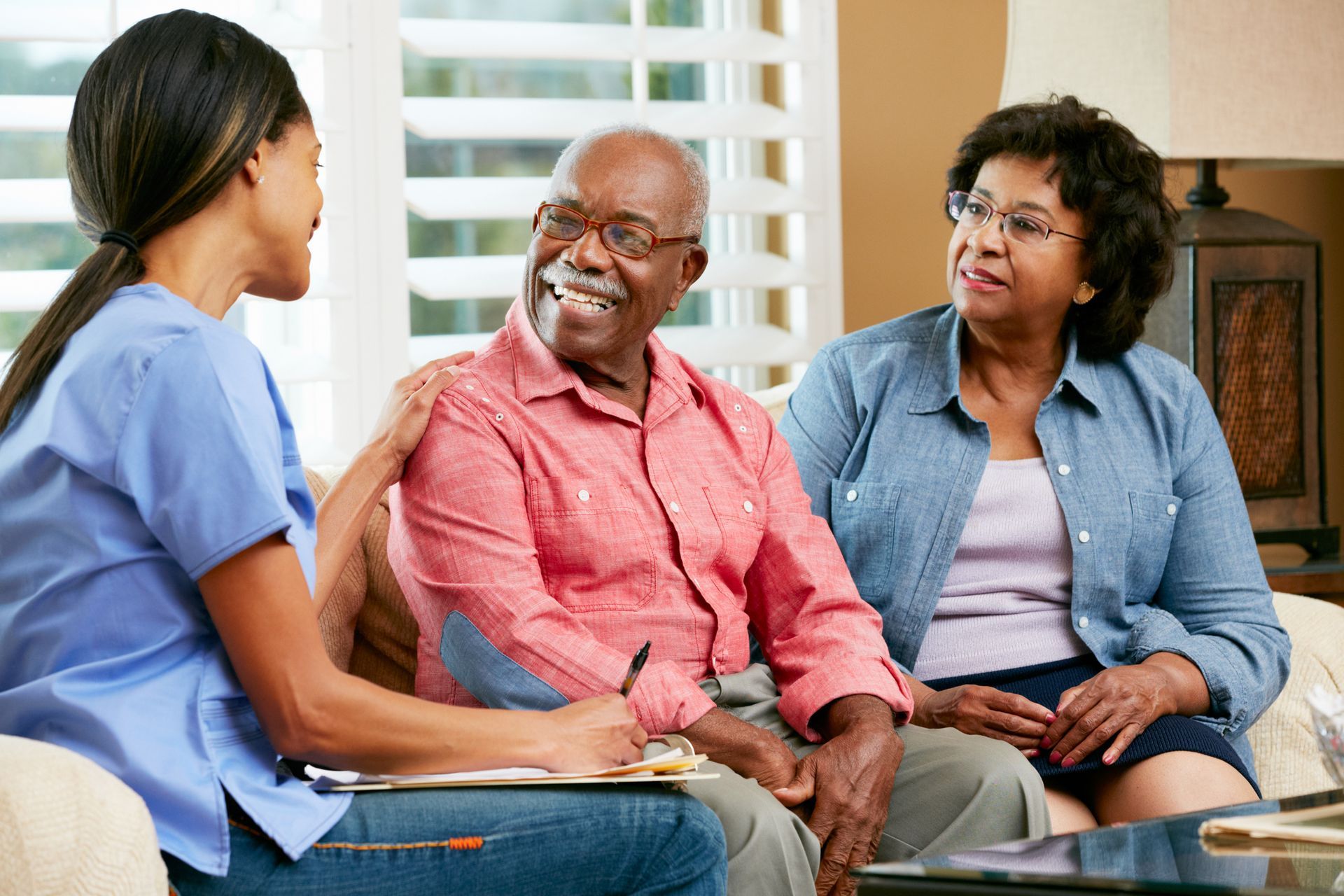 A nurse in blue scrubs consults with an elderly couple on a sofa; smiling, indoor setting.