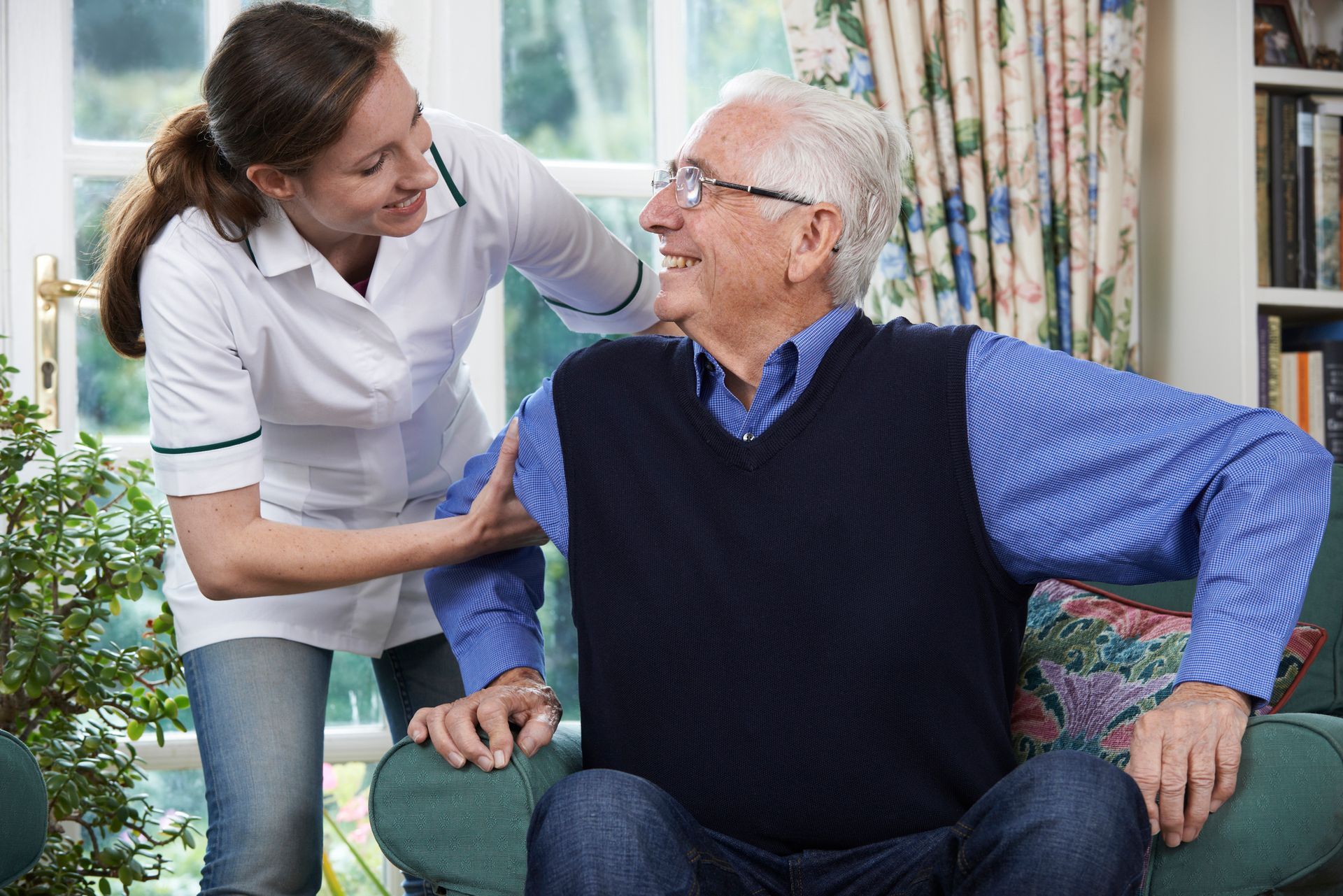 Nurse assisting an elderly man to stand from a chair. They are in a living room.