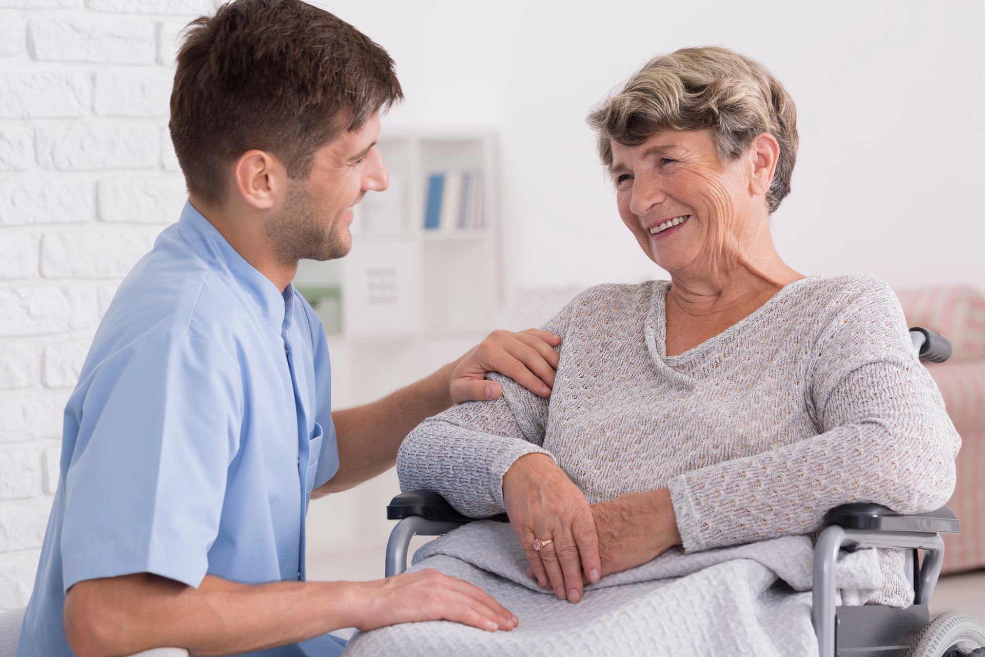 Caregiver smiles at an elderly woman in a wheelchair, indoors.