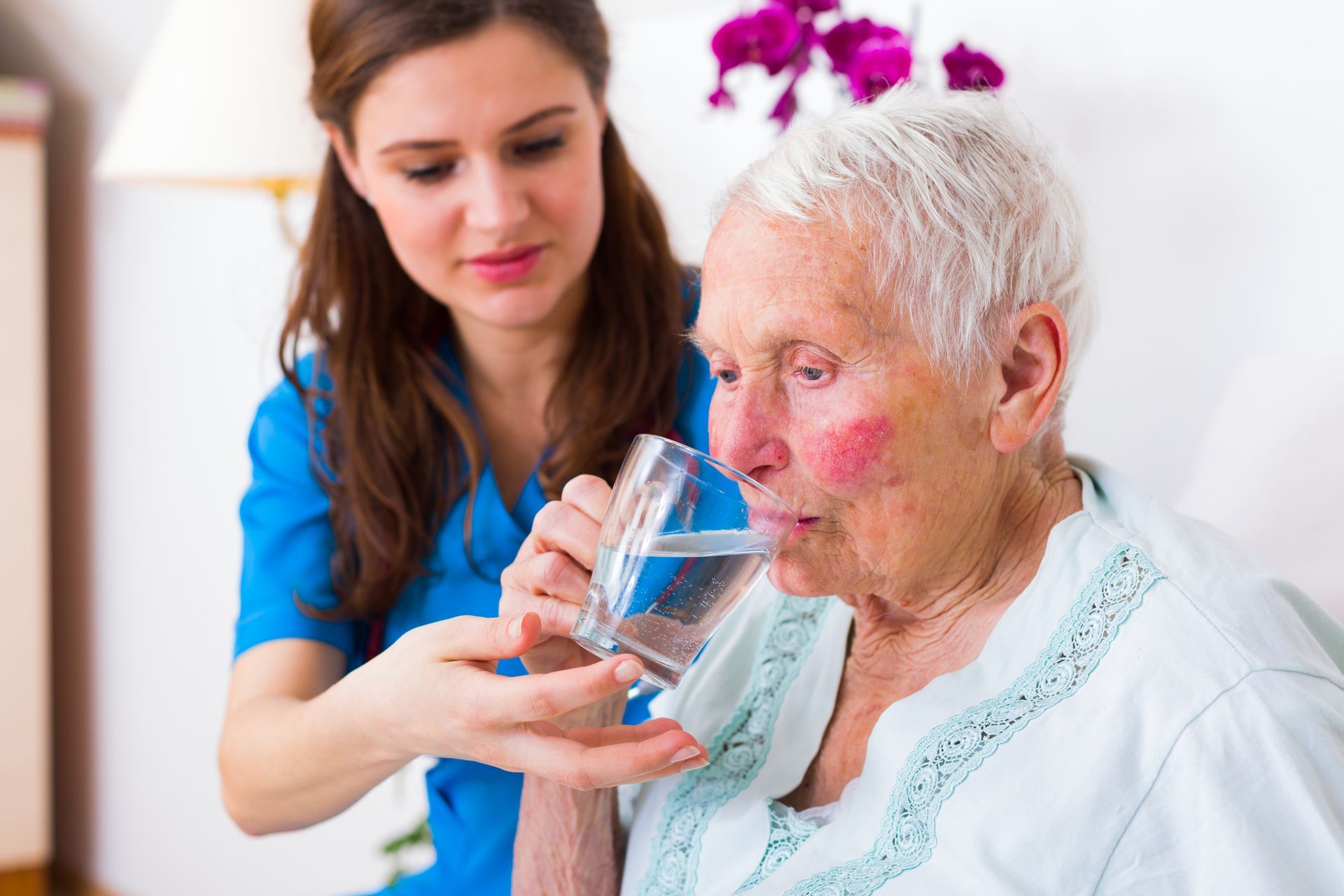 Caregiver assisting elderly woman with a drink of water. Indoor setting; blue and white.
