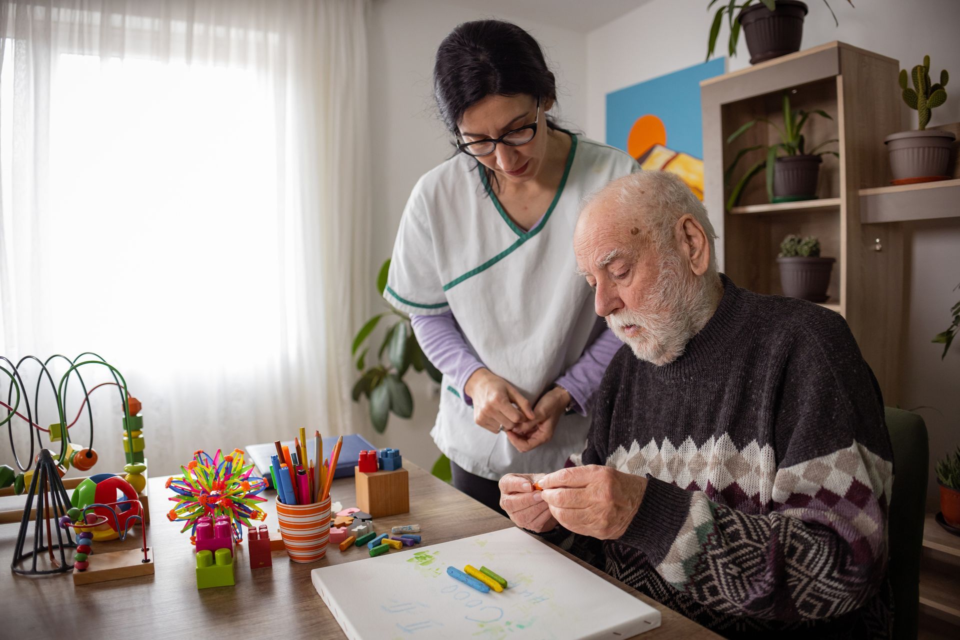 Caregiver assisting senior man with art therapy using crayons at a table.