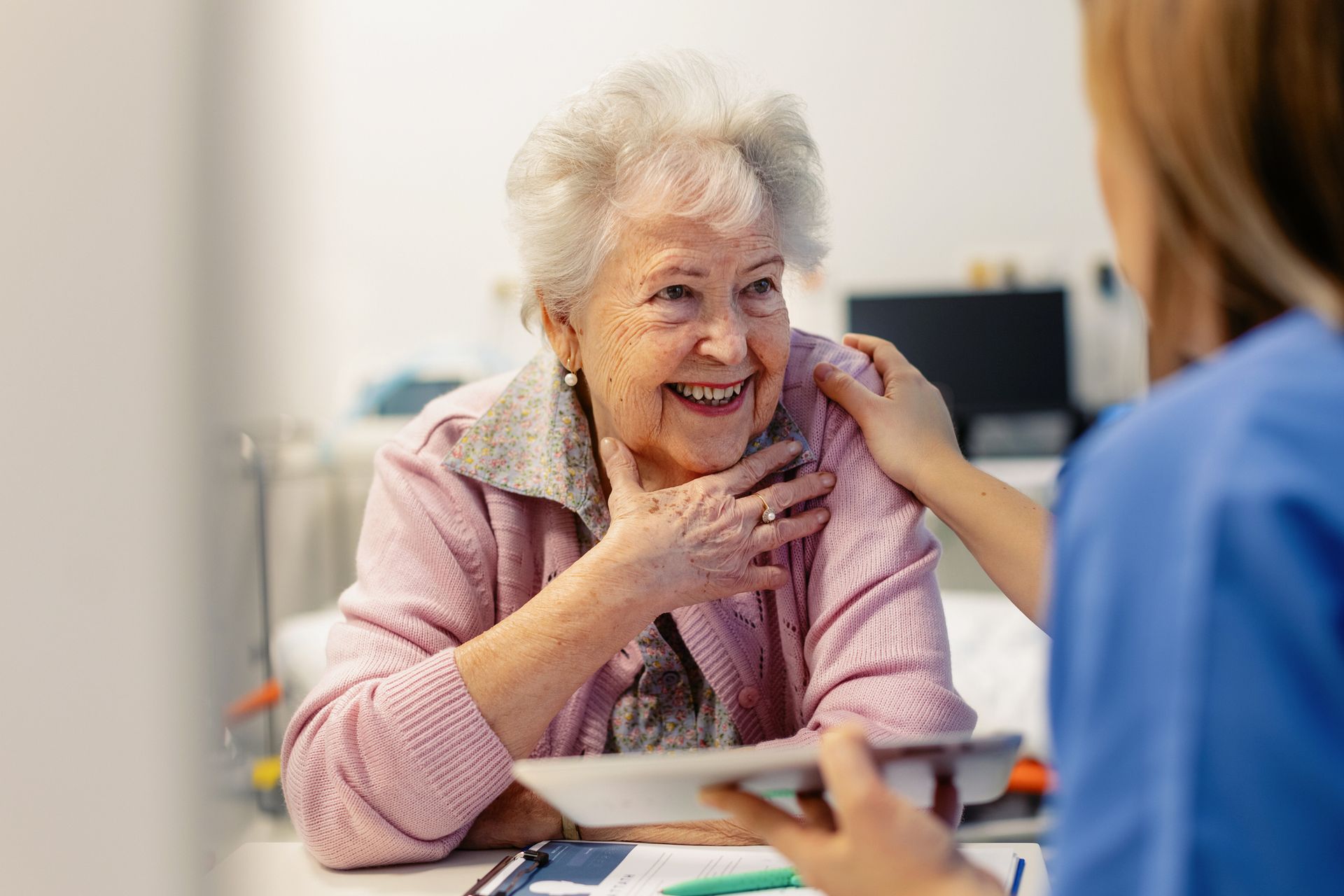 Elderly woman smiles, touched on arm by a nurse, indoors.