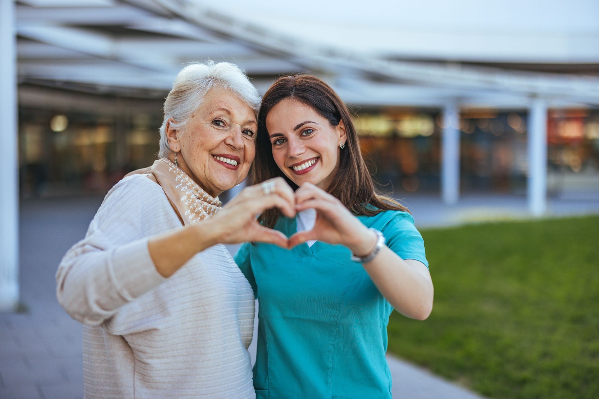 An older woman and a younger woman forming a heart with their hands, smiling outside.