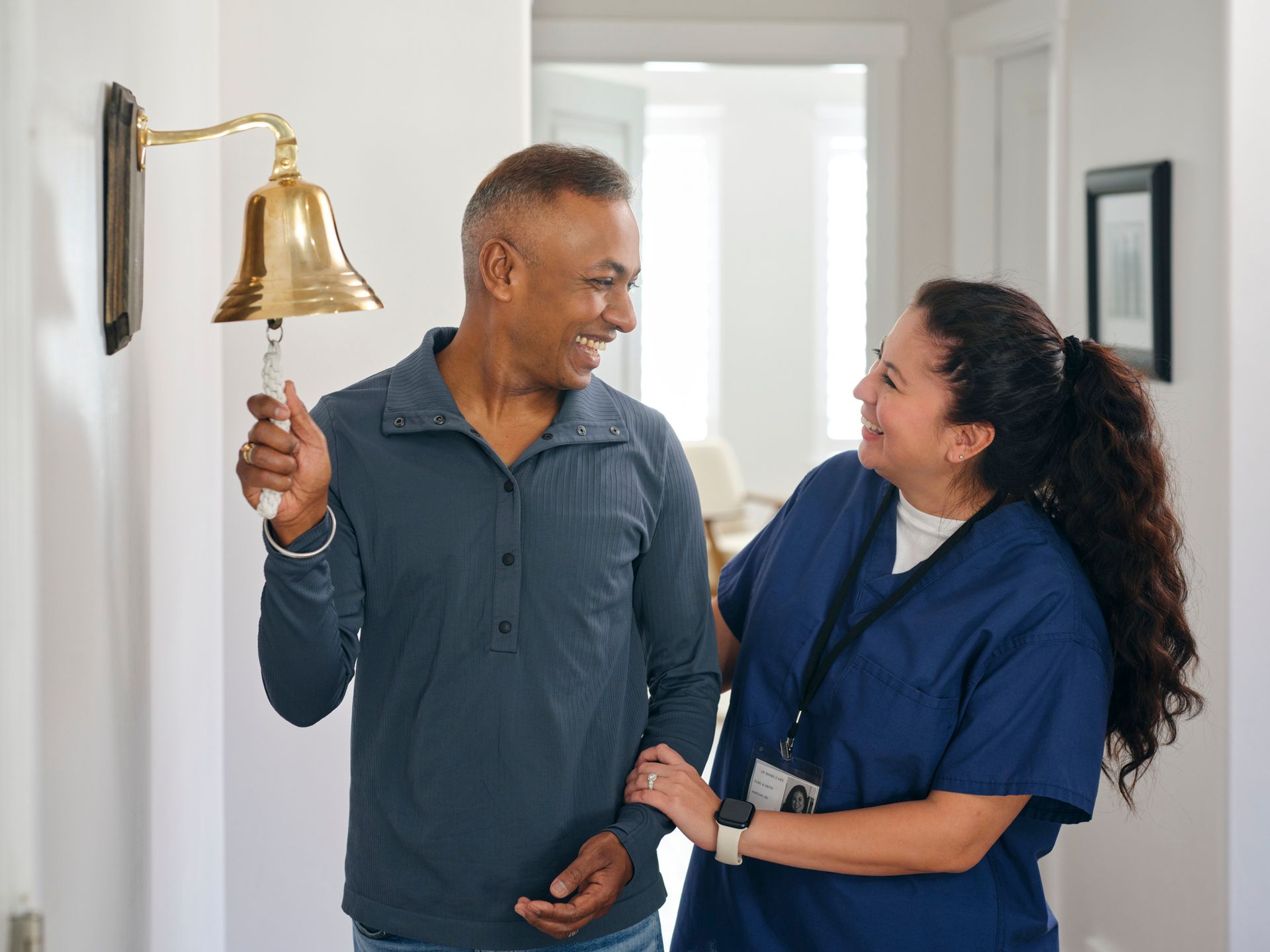 Man ringing a gold bell, smiling at a nurse. They are in a well-lit hallway.