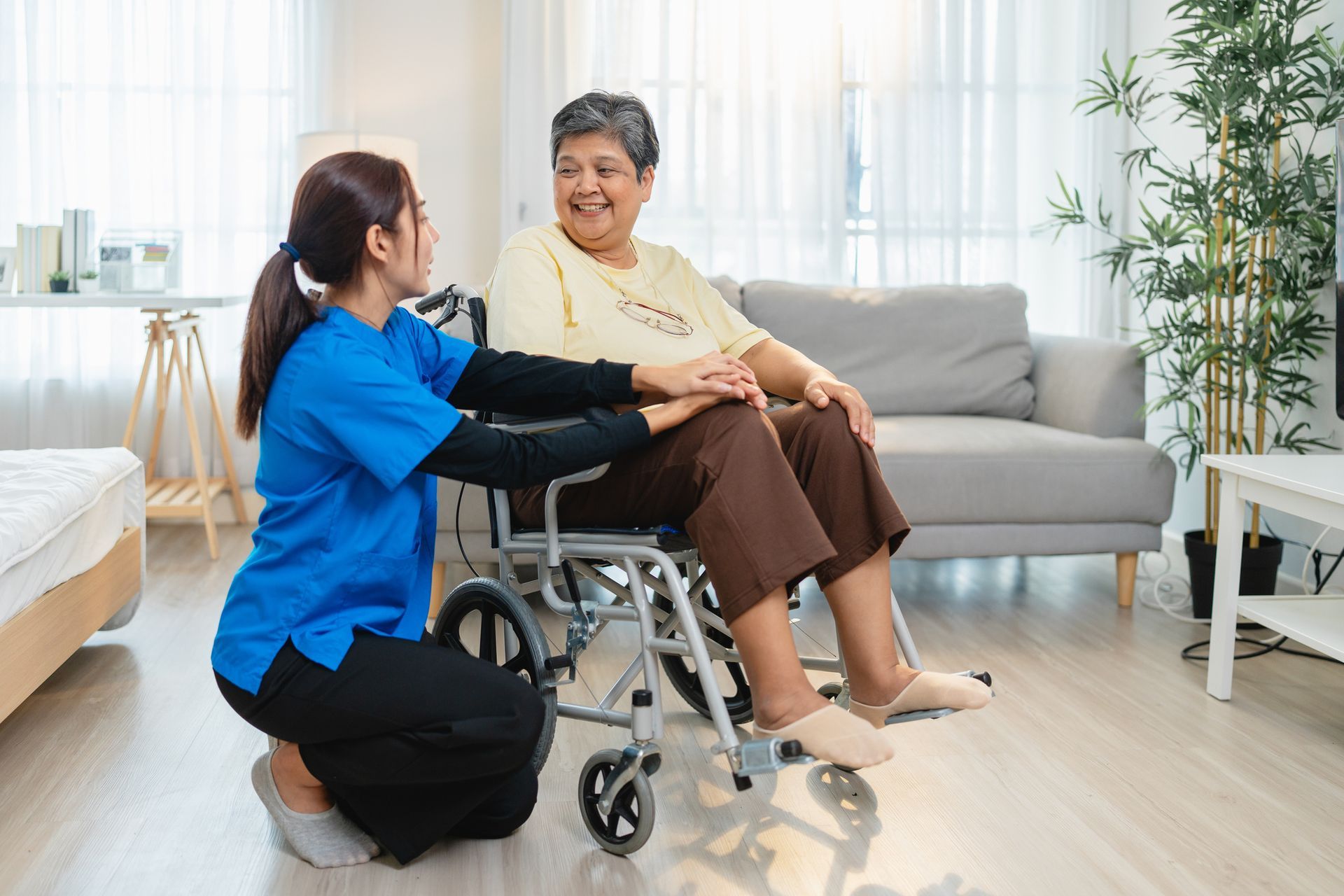 Caregiver kneels, holding hands with senior woman in wheelchair, indoors, smiling.