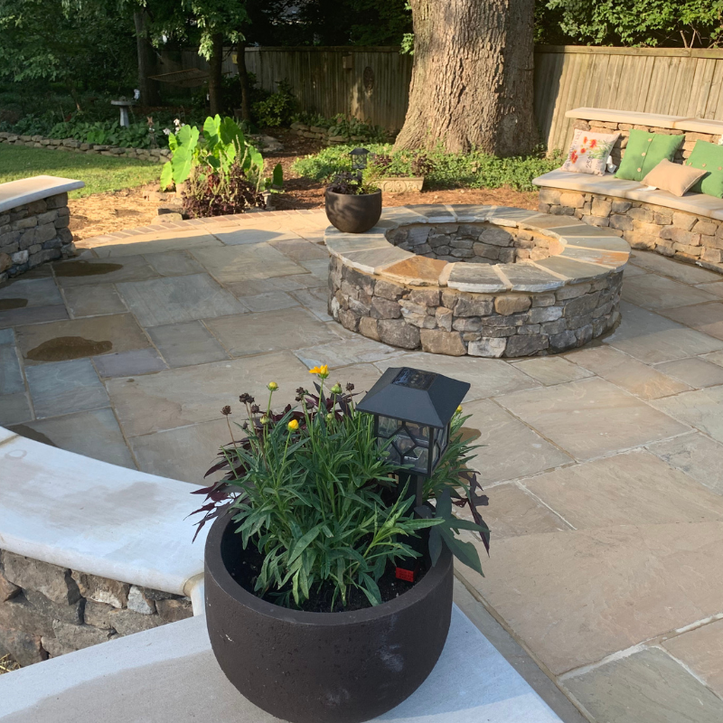 Stone patio with fire pit, built-in bench, and potted plants. Sunny setting with a tree.
