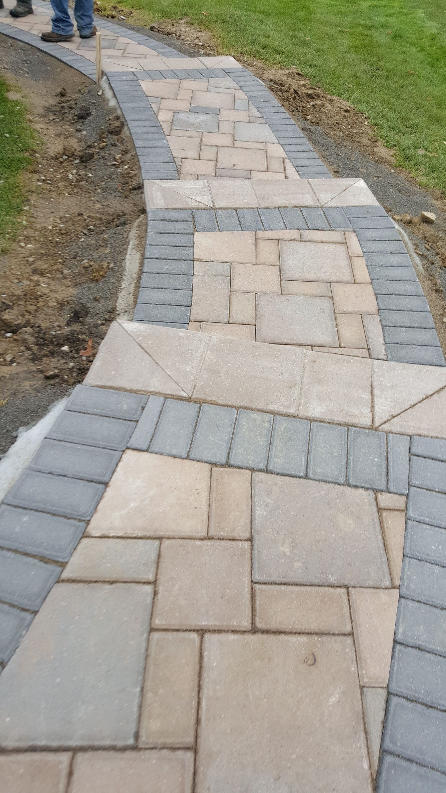 A paved walkway features a central pattern of rectangular beige stones bordered by a row of dark grey rectangular pavers.
