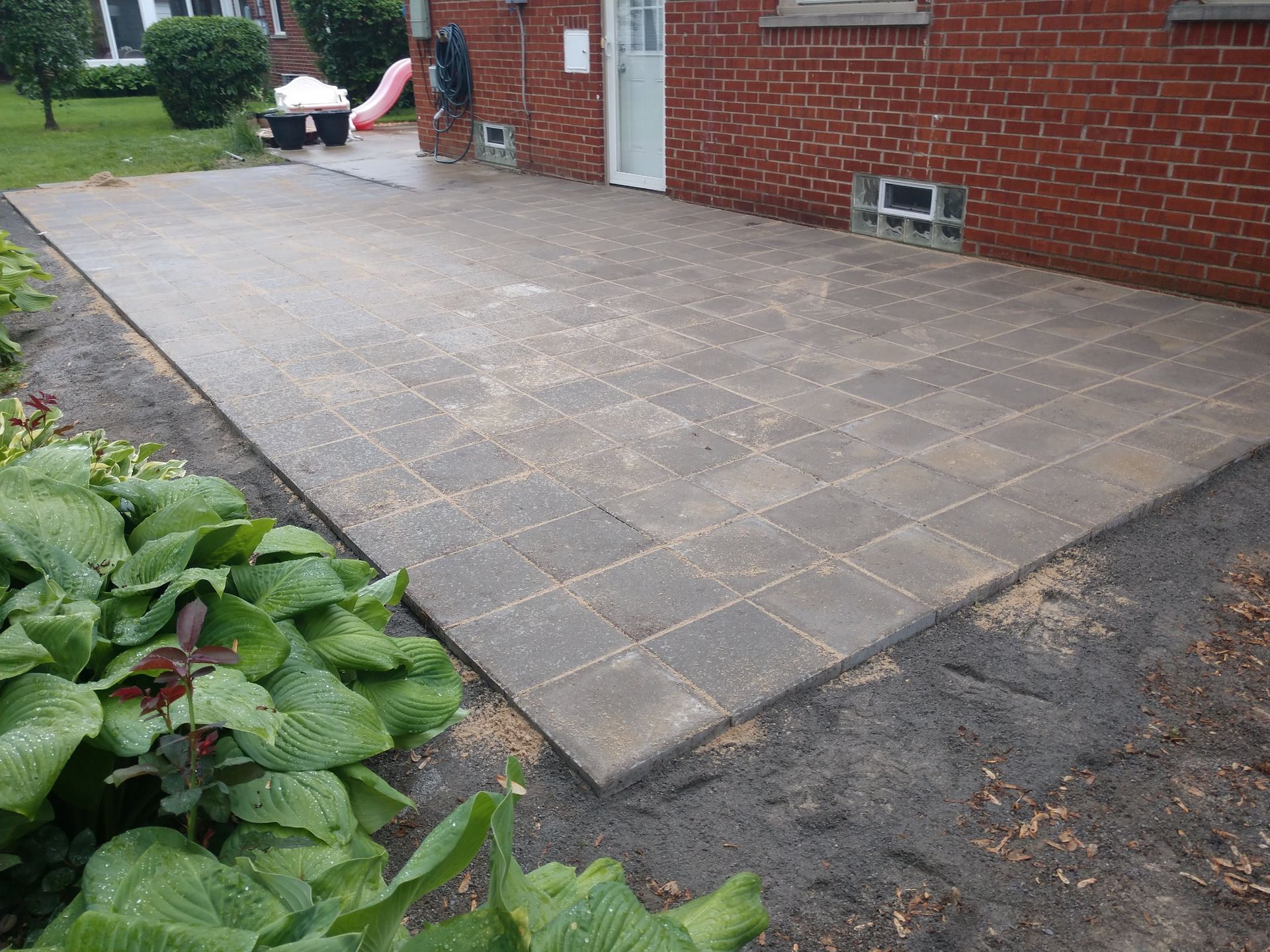 A newly installed rectangular patio made of square gray pavers sits adjacent to a red brick house with glass block vents.