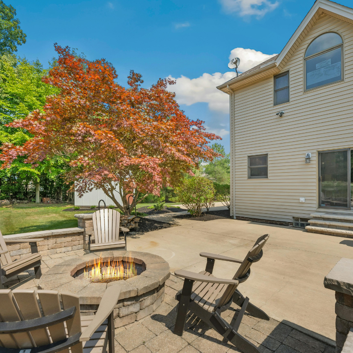A stone fire pit with a lit fire surrounded by patio chairs on a concrete patio beside a beige house and an autumn tree.