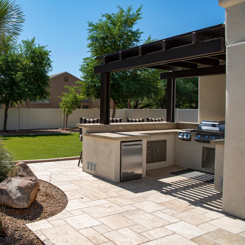 Outdoor kitchen with grill, fridge, and countertop under a wooden pergola on a patio.
