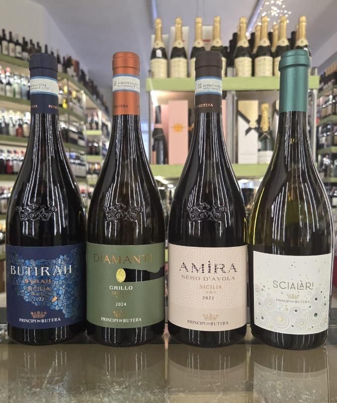 Quattro bottiglie di vino con etichette colorate esposte in un negozio con scaffali di bottiglie sullo sfondo.