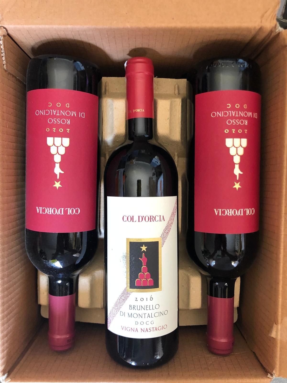 Tre bottiglie di vino rosso in una scatola di cartone, due con etichette rosse e una con etichetta bianca.