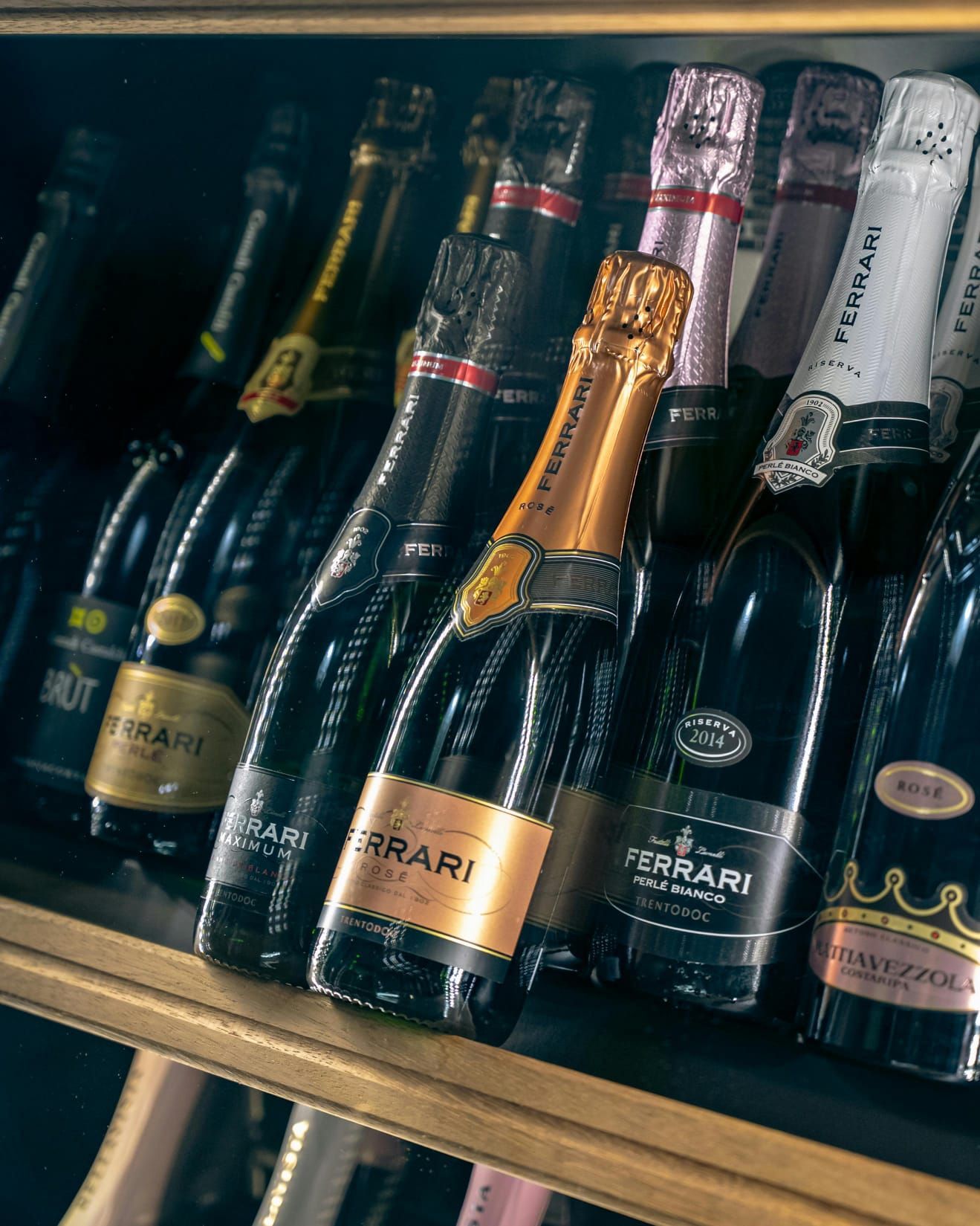 Bottiglie di spumante su uno scaffale; etichette dorate, nere e bianche; varie forme e dimensioni.
