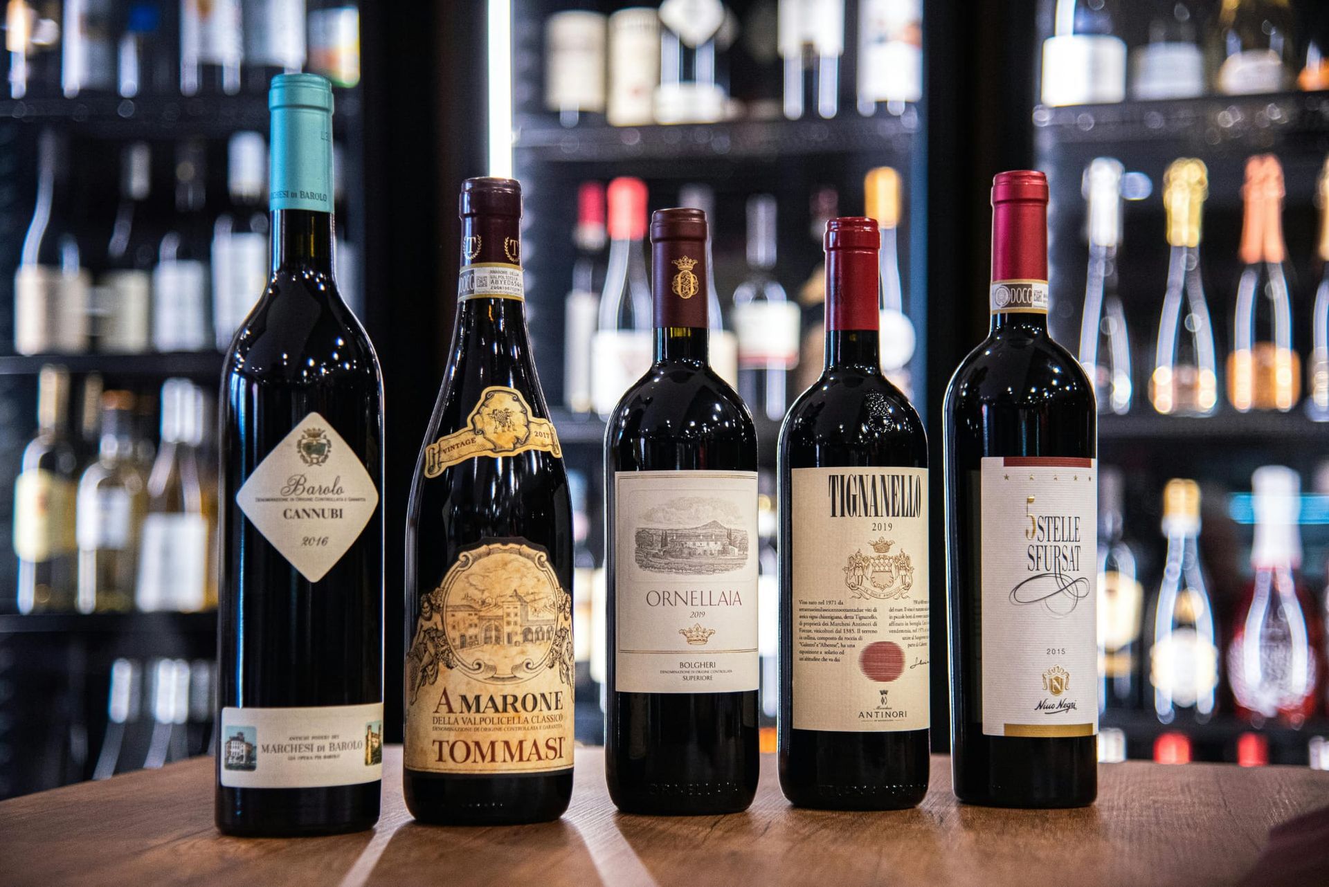 Cinque bottiglie di vino su un tavolo con uno scaffale per bottiglie sullo sfondo. Bottiglie rosse e scure con etichette colorate.