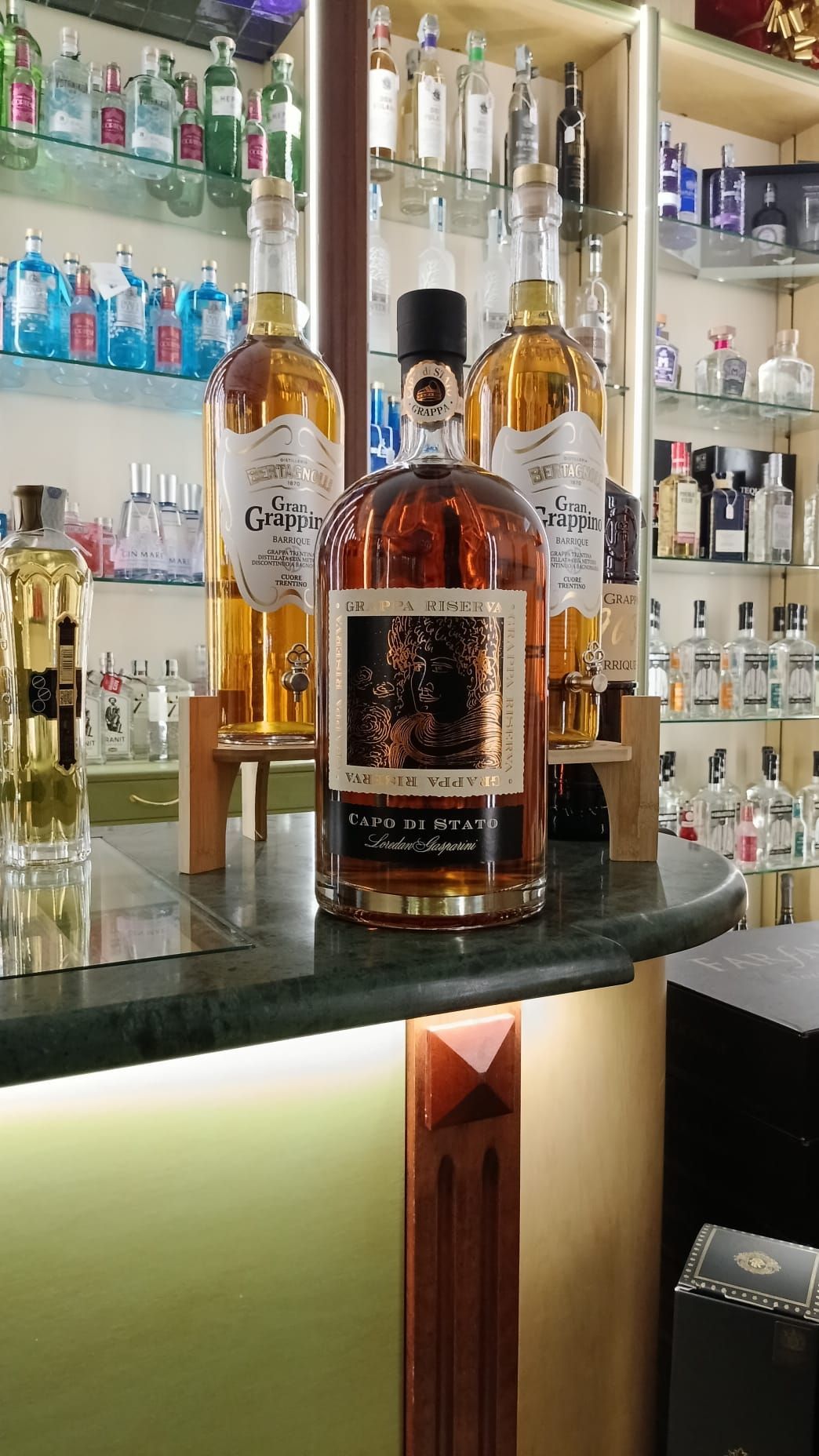 Bottiglia di liquore color ambra su un bancone, con due bottiglie simili dietro. Ambiente da negozio di liquori.