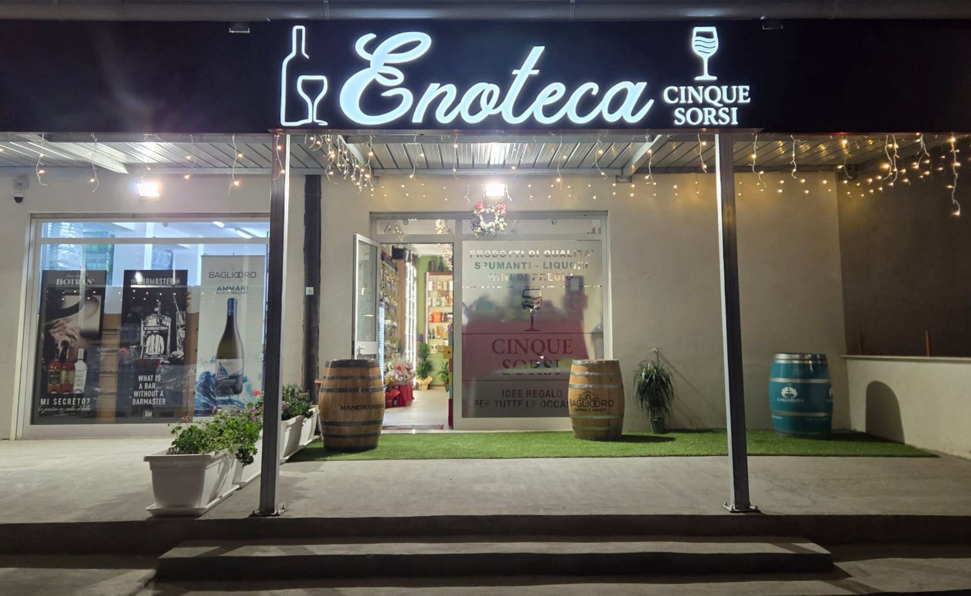 La vetrina dell'Enoteca Cinque Sorsi di notte. Insegna illuminata, due botti di vino e prato sintetico verde.