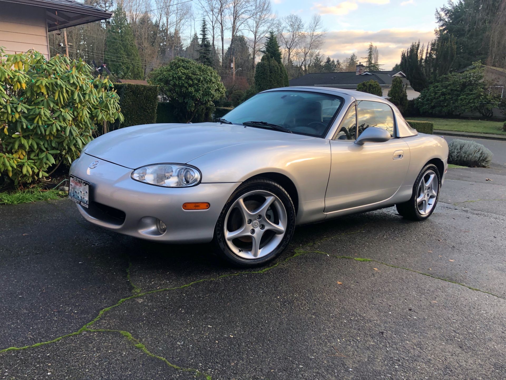 Silver Mazda Miata sports car on a wet driveway, trees in the background.