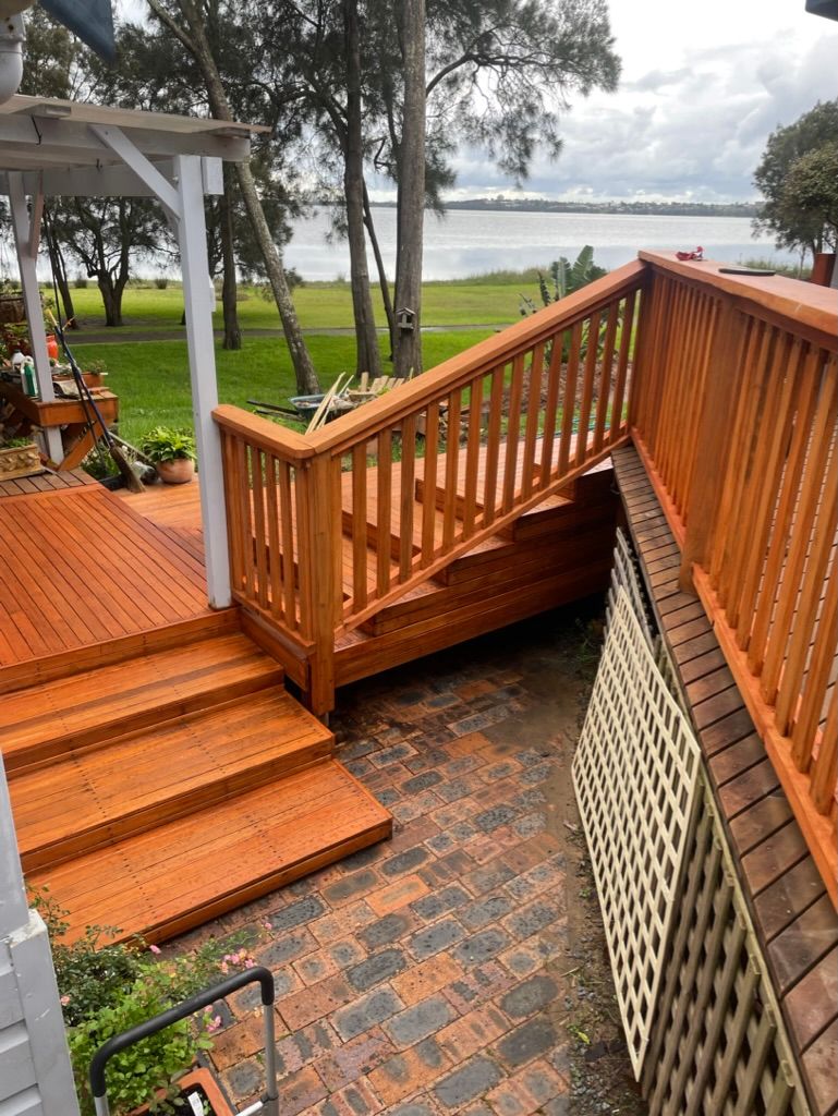 Wooden deck and stairs leading down to a brick patio, overlooking a body of water and grassy area. — Gary Eslick Home Improvements In Berkeley Vale, NSW
