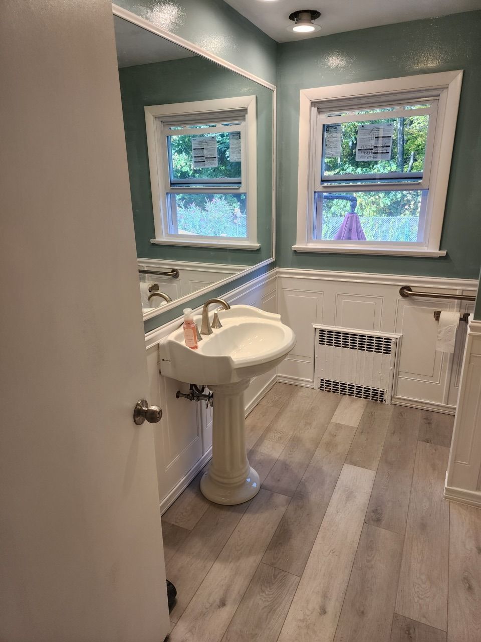 A bathroom with a sink , mirror , and two windows.