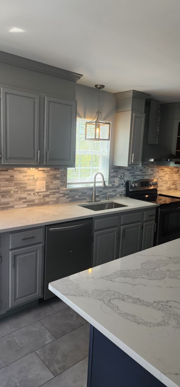 A kitchen with gray cabinets , white counter tops , black appliances and a large island.