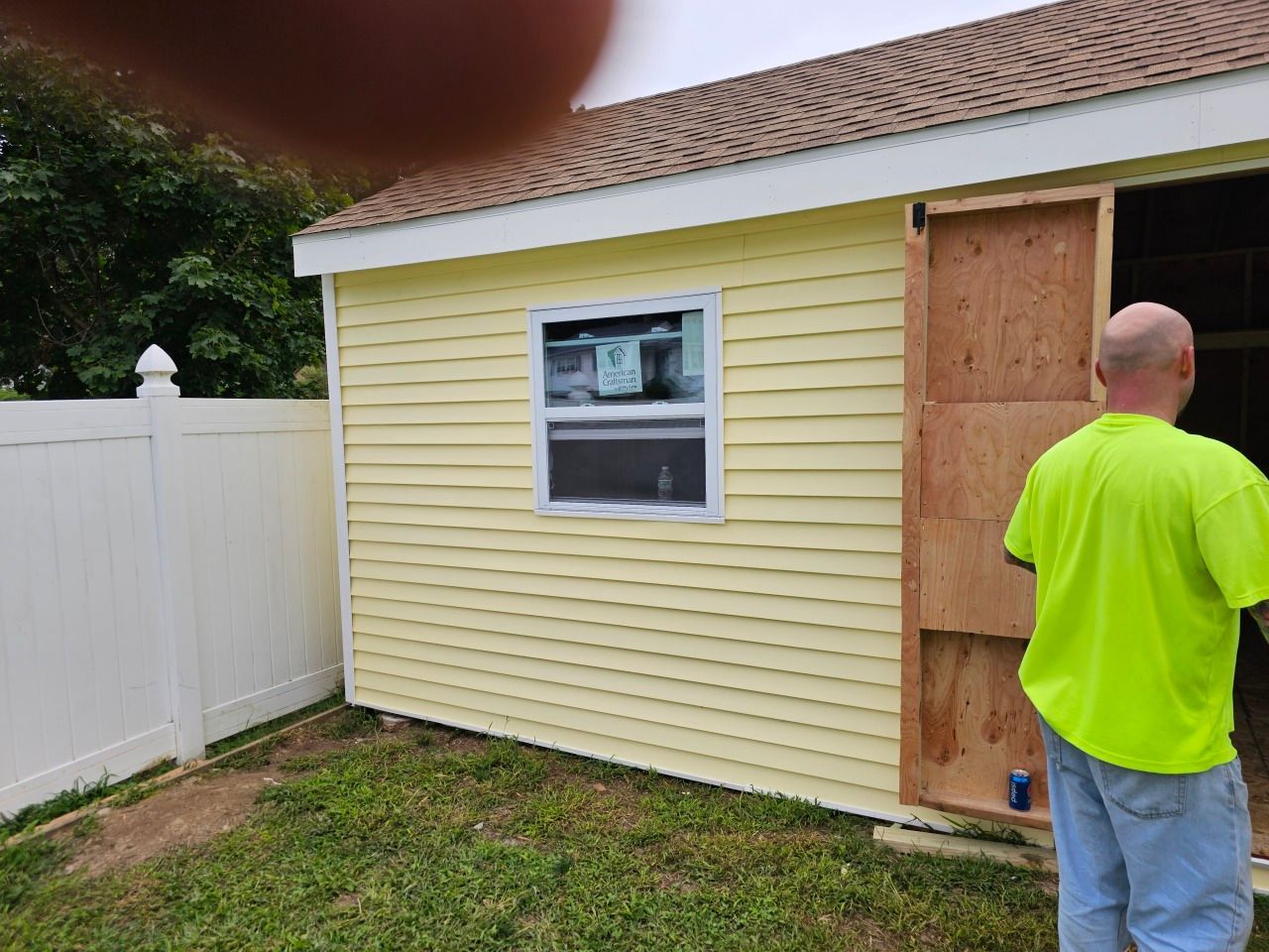 A man in a neon green shirt is standing in front of a yellow shed.