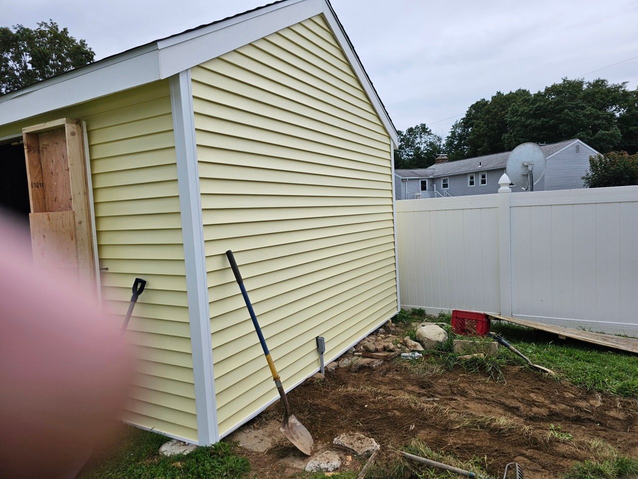 A yellow shed with a white fence and a shovel in front of it.