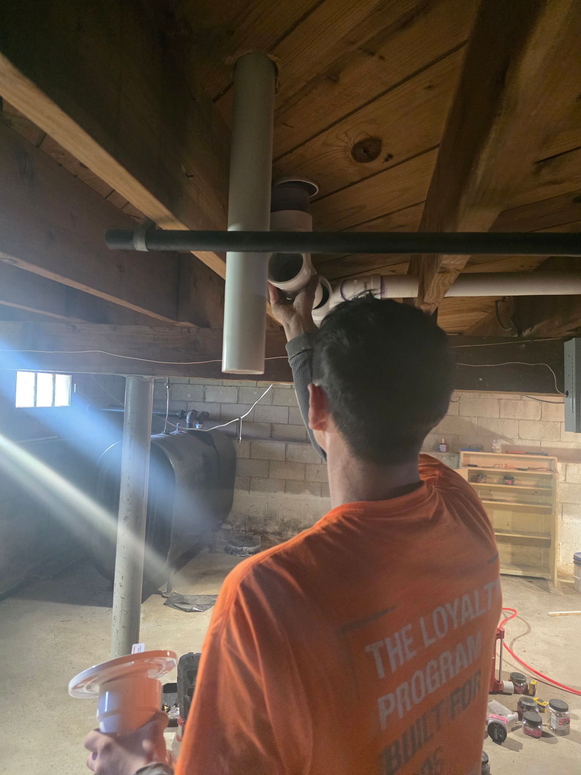 A man in an orange shirt is working on a pipe in a basement.