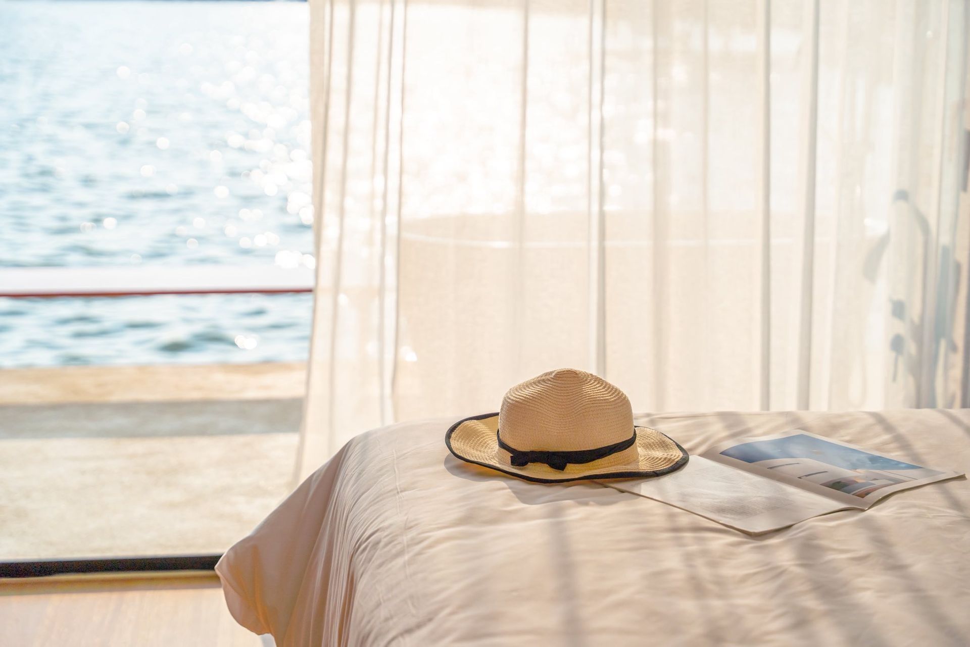 Straw hat on a bed by a sheer-curtained window overlooking the sea