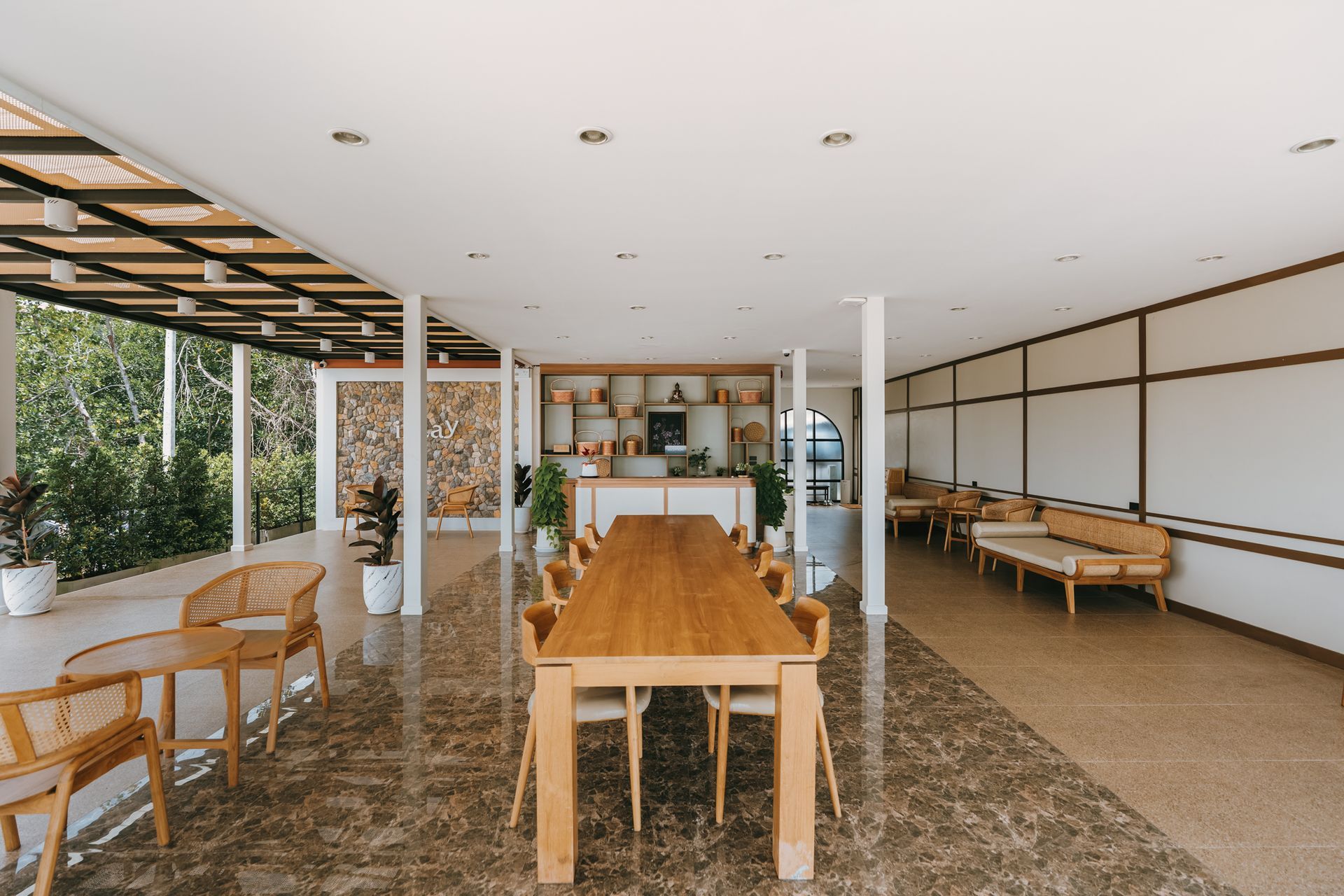 Open-air café with wooden tables and chairs, white columns, and a long communal table on a stone floor.