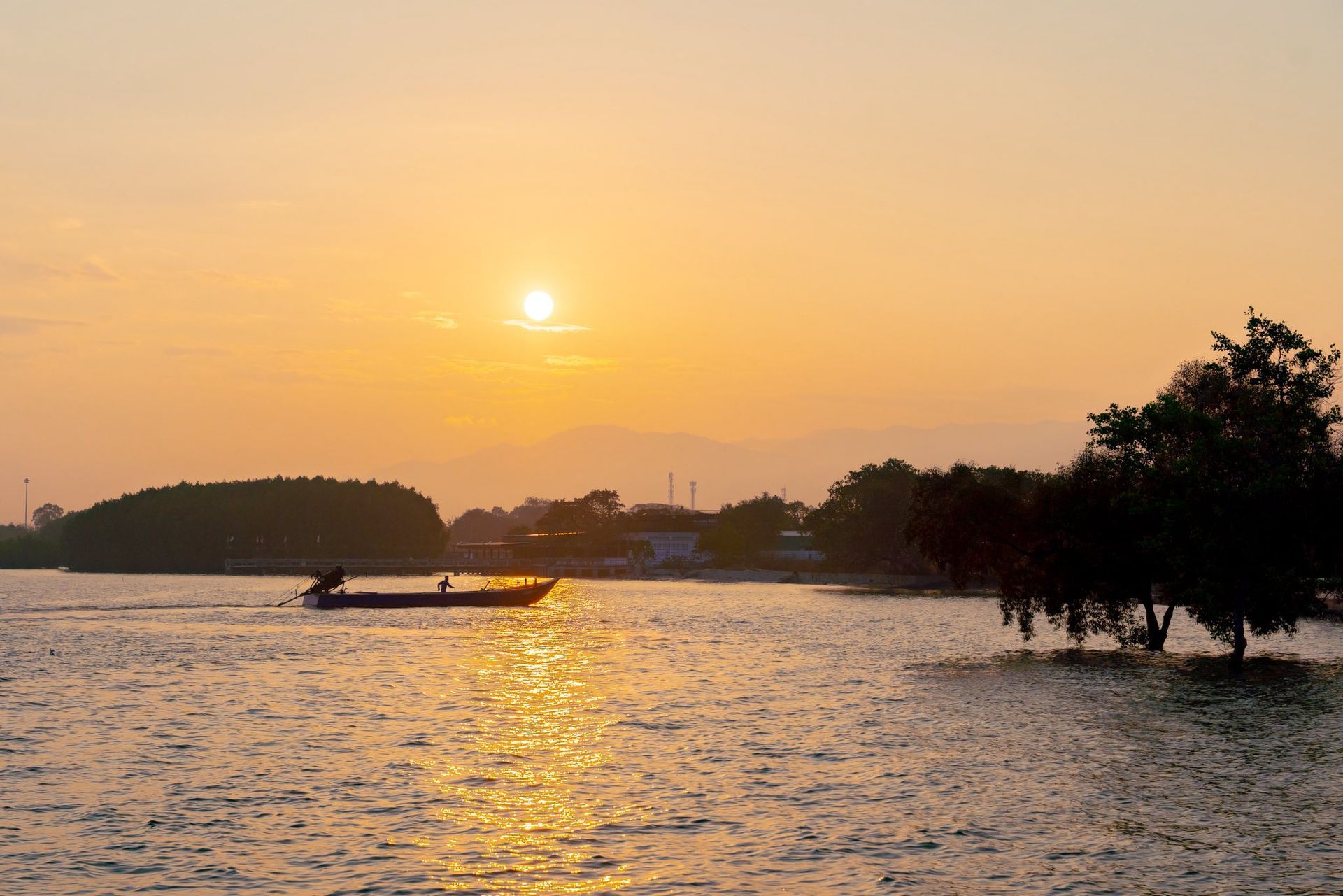 Golden sunset over calm water with a small boat, distant shoreline, and silhouetted trees