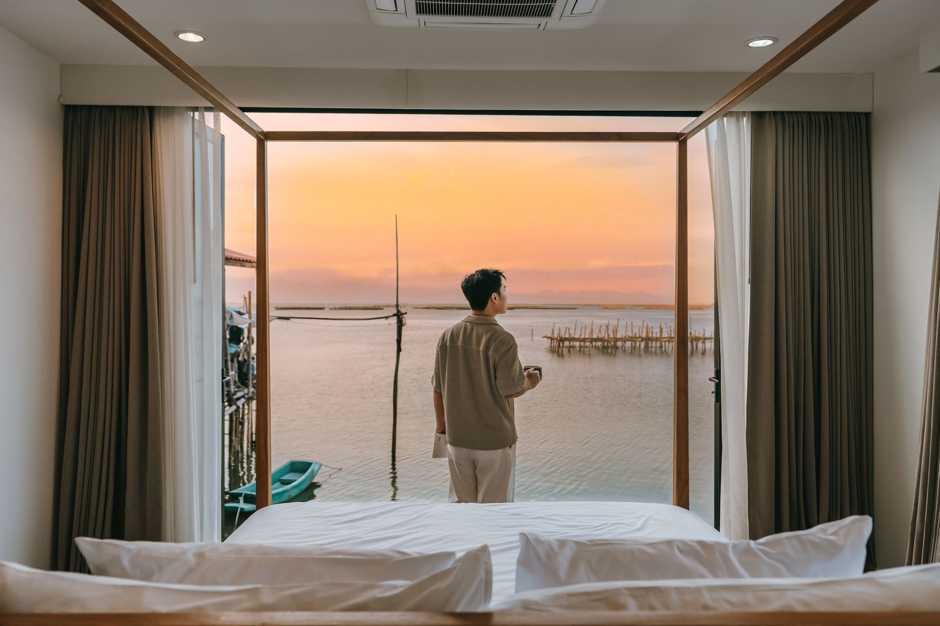 Person standing by an open window in a bedroom, looking out at a sunset over the water.
