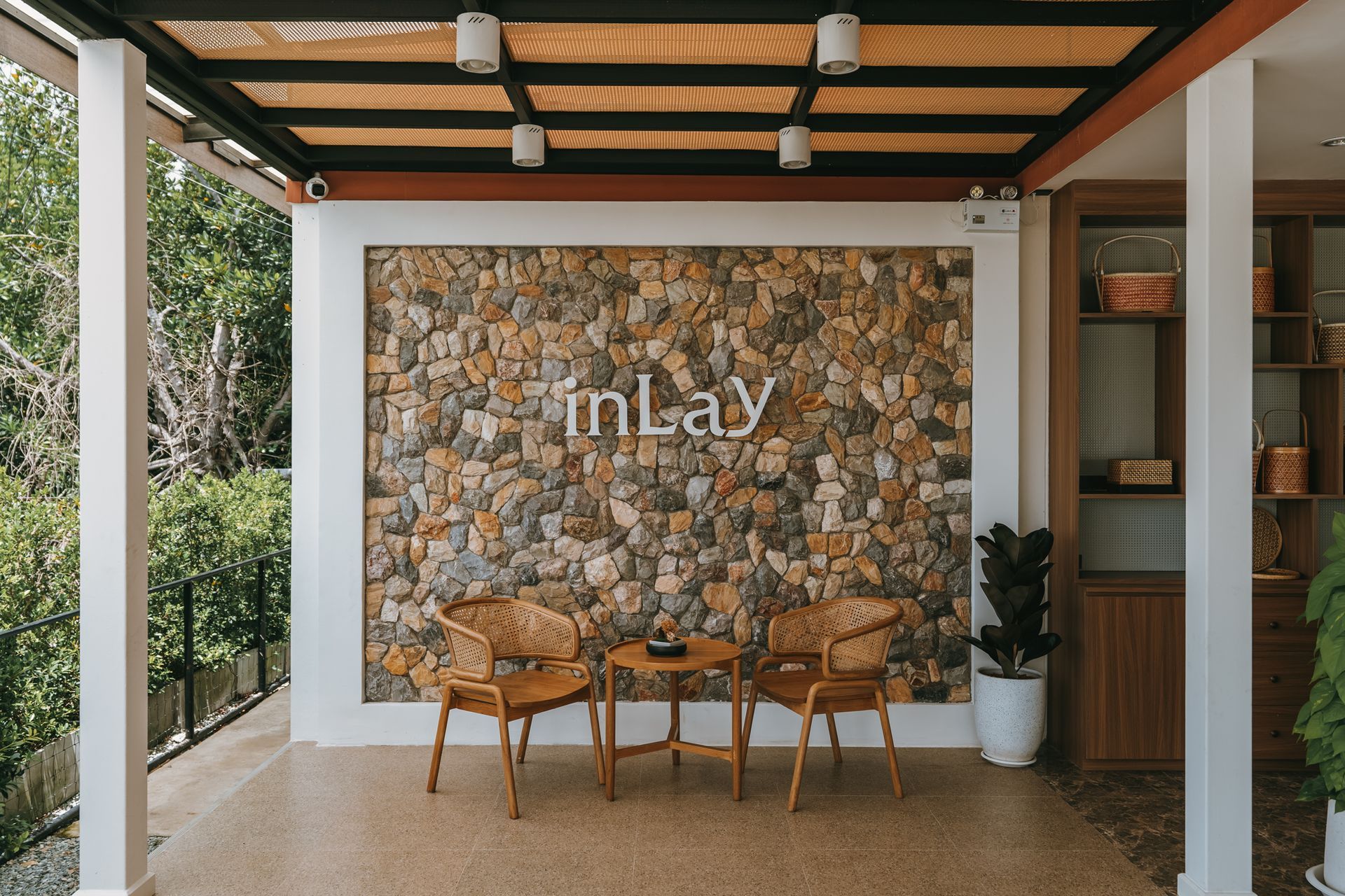 Outdoor café patio with stone wall, wooden chairs, and potted plants under a pergola