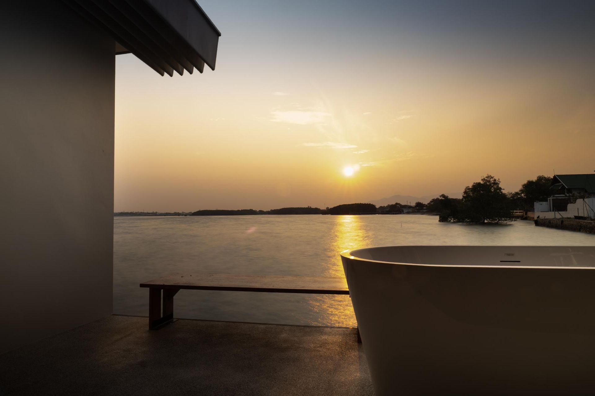 Sunset over calm water viewed from a patio, with a bench and silhouetted shoreline.