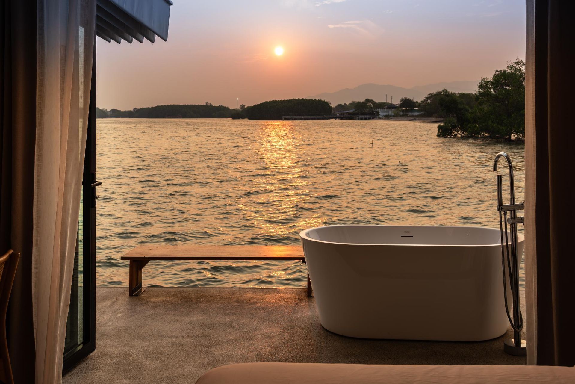 Sunset over a calm lake with a deck, freestanding tub, and bench beside open curtains