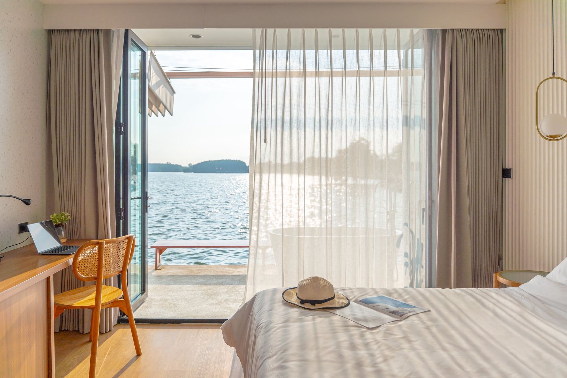 Bright seaside bedroom with open balcony doors, sheer curtains, desk, and a bed facing the ocean.