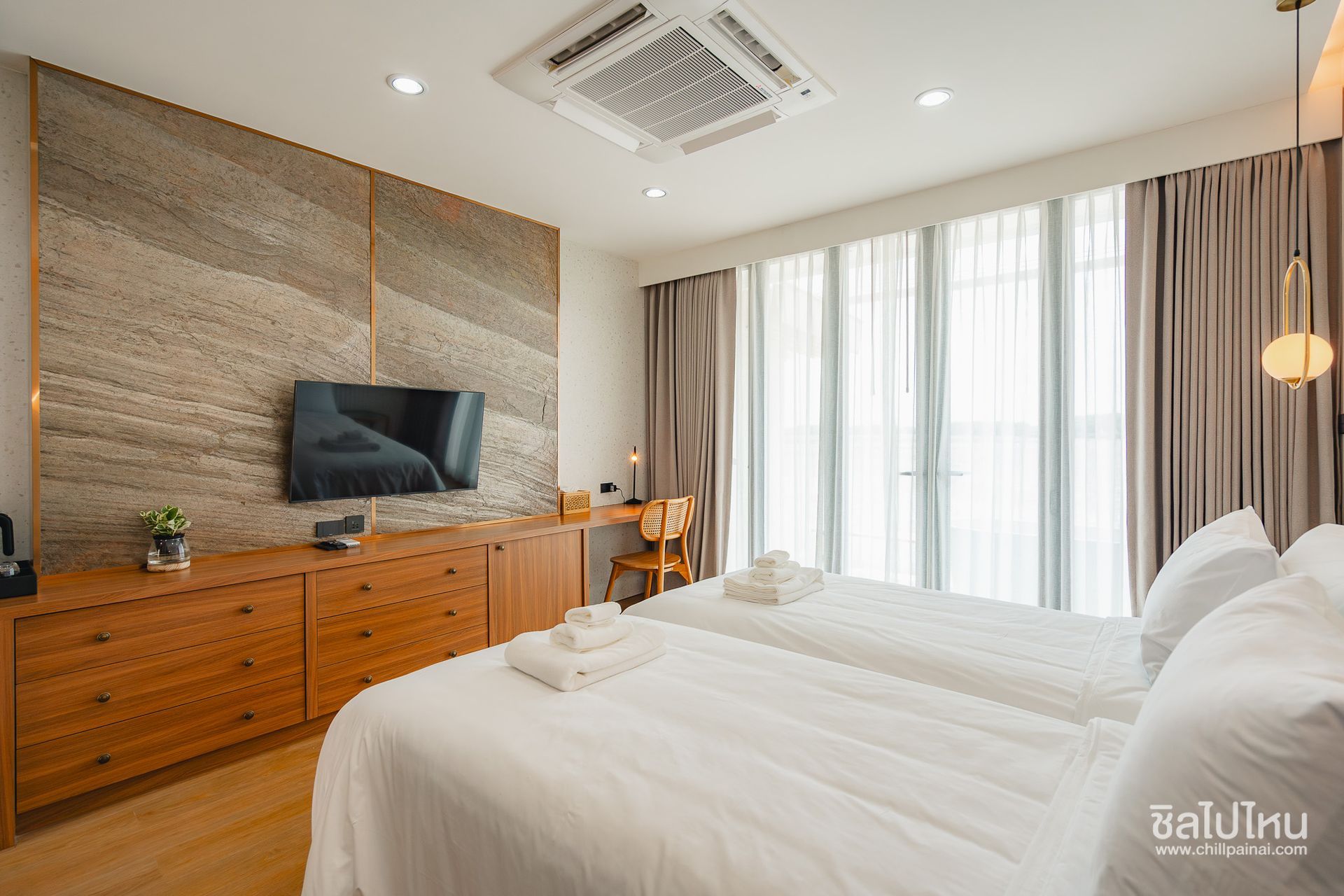Bright modern bedroom with a large bed, wooden dresser, wall-mounted TV, and floor-to-ceiling curtains