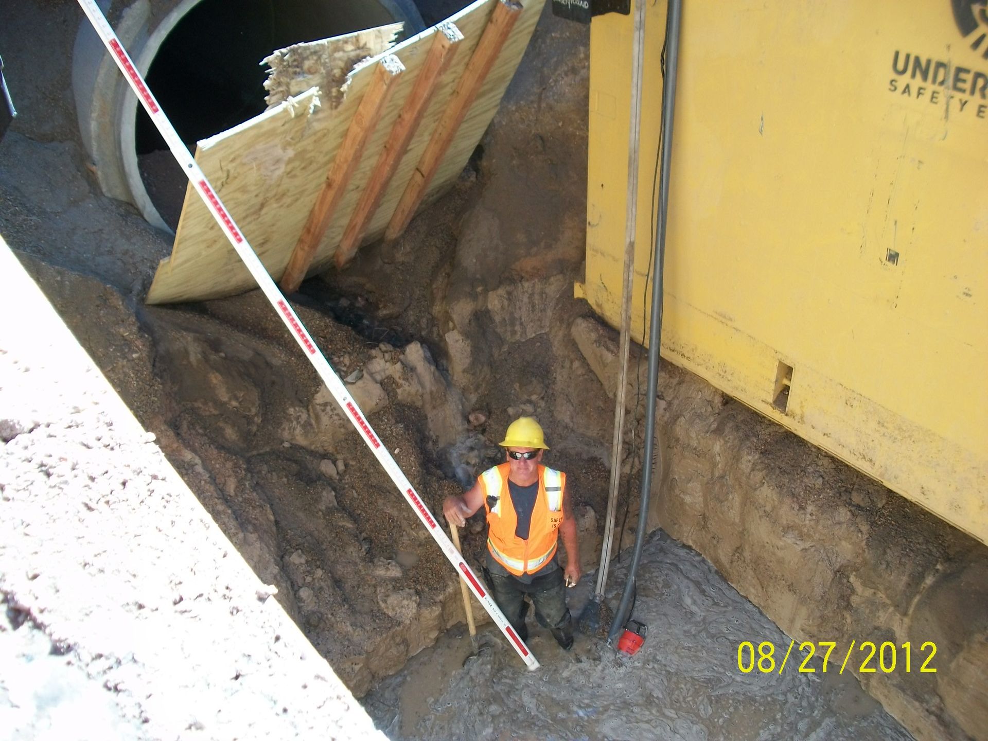 Worker in hard hat and orange vest standing in a deep construction trench beside a yellow wall and safety tape